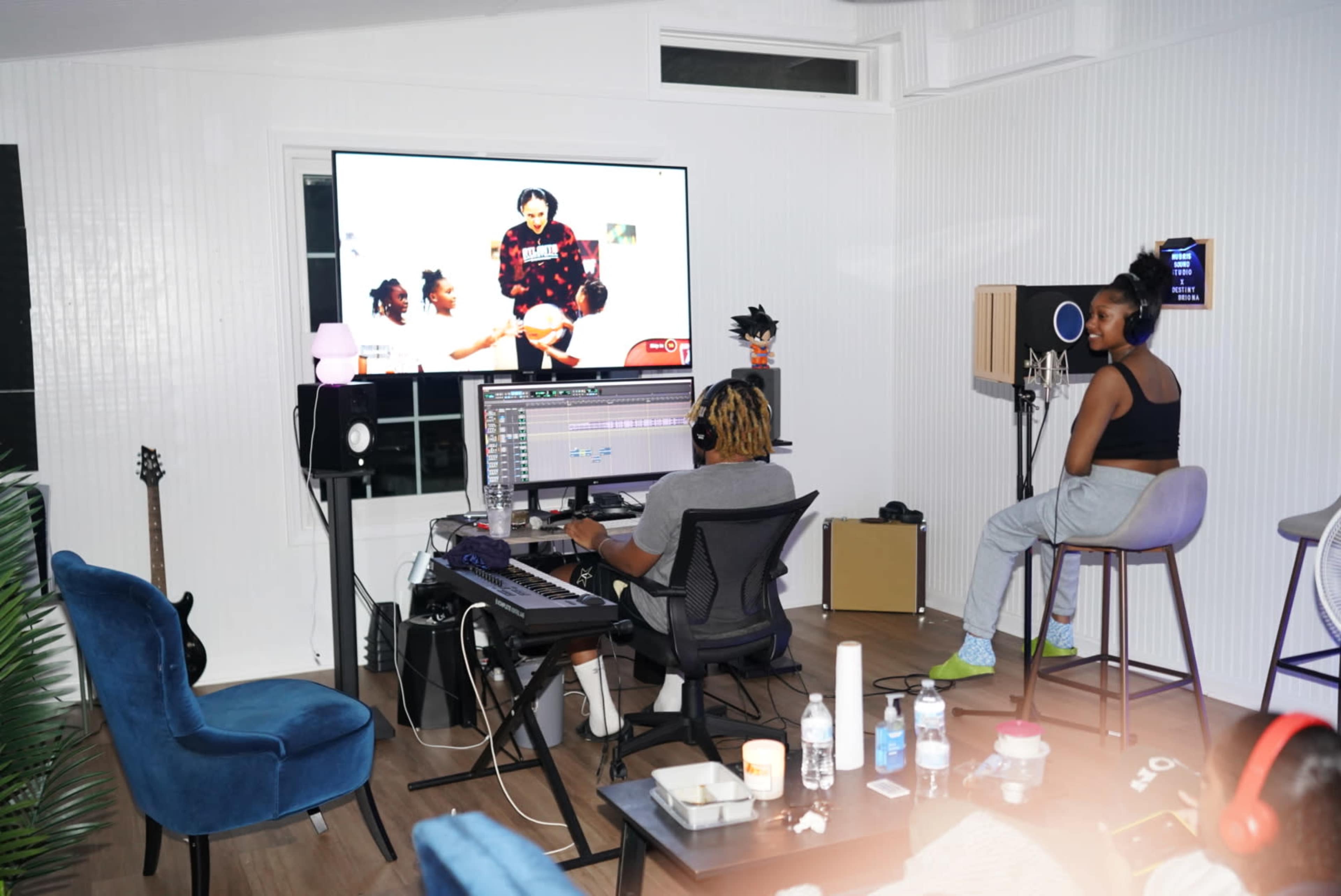 Two individuals are seated in a music studio, one working on a computer with music software, while the other listens through headphones, with a large screen displaying a video.