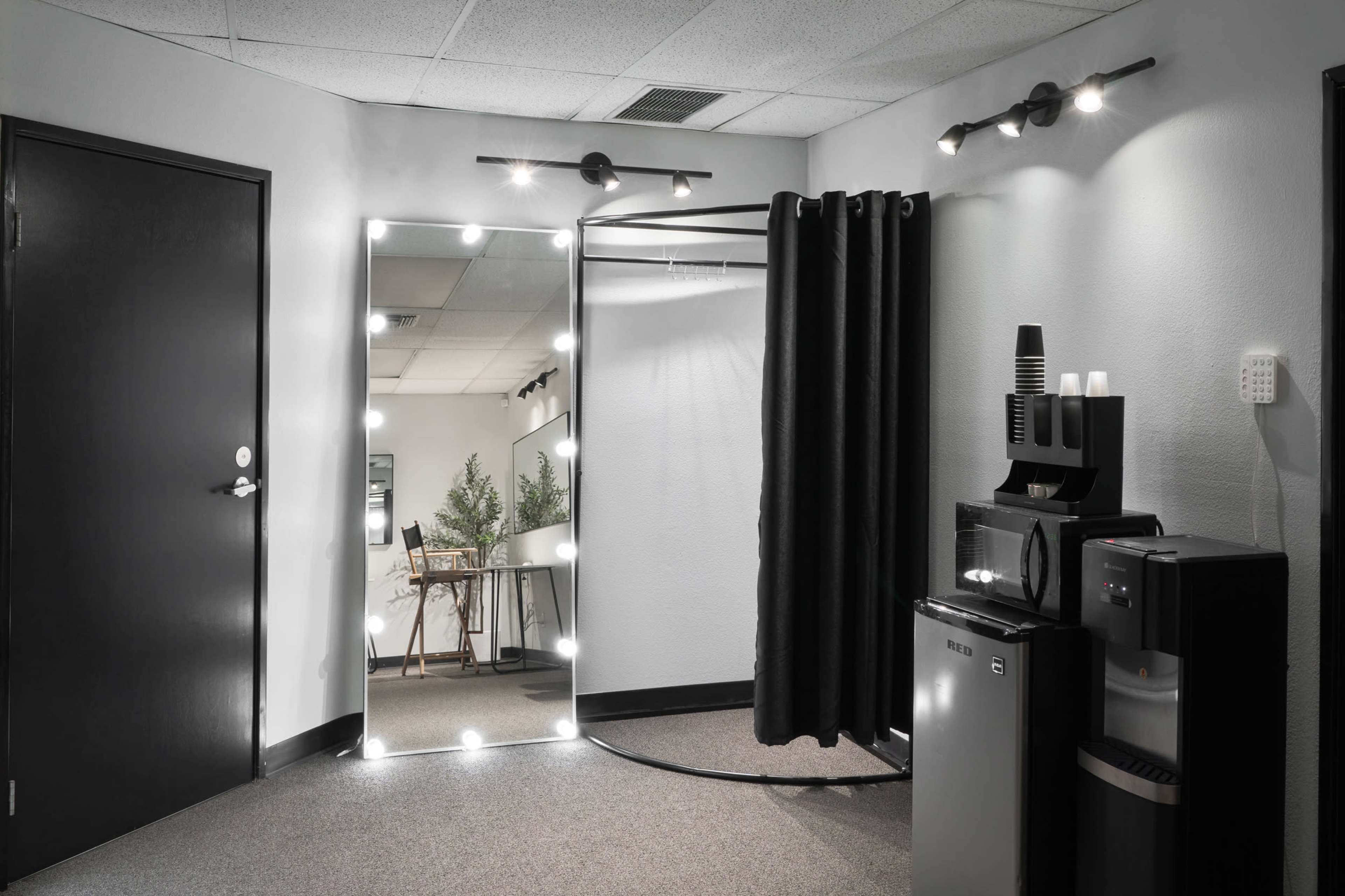 A well-lit room featuring a large mirror with lights, a curved mirror, a black curtain, and various appliances including a microwave and a water cooler.