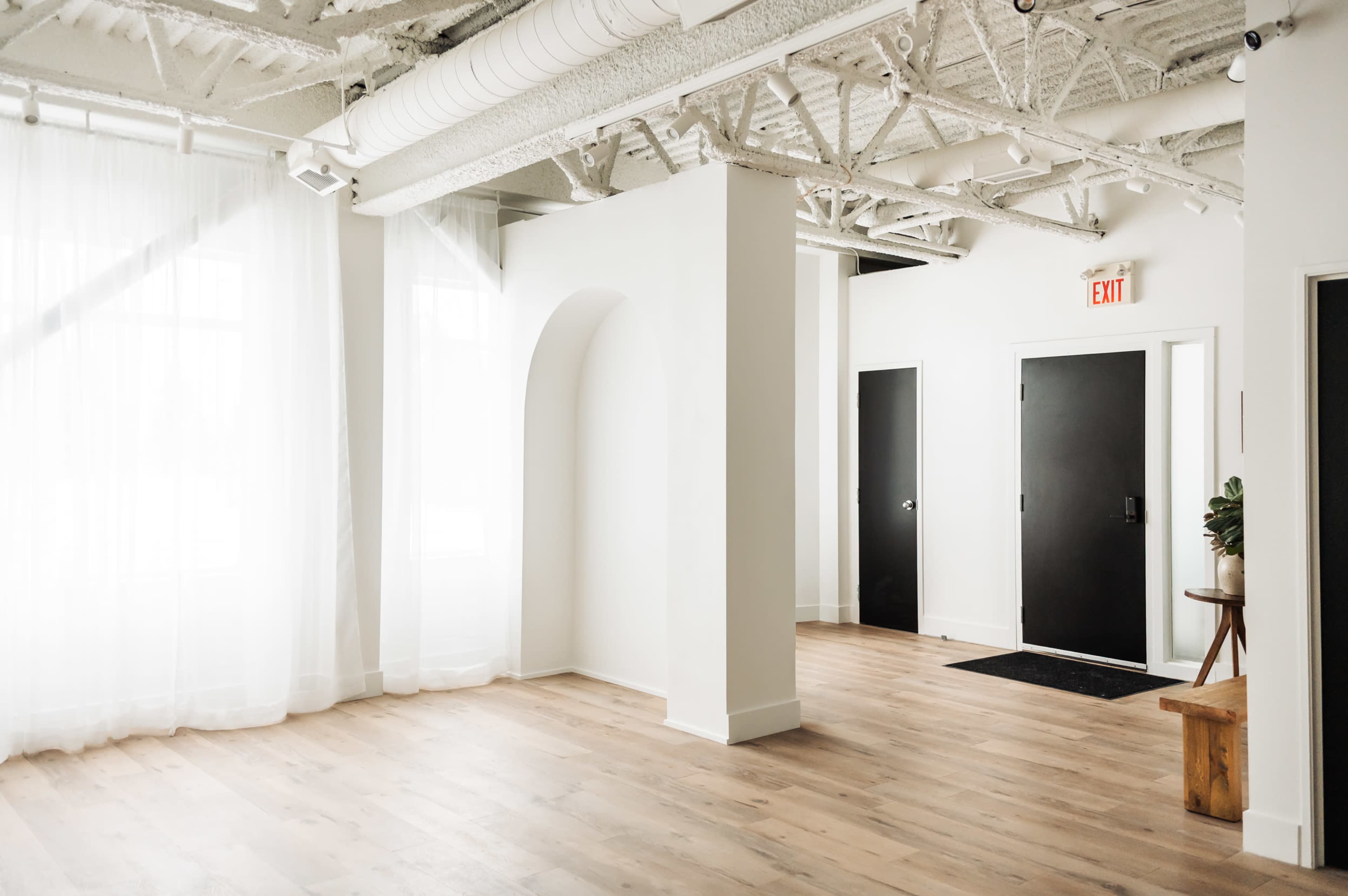 The image shows a spacious, airy room with hardwood floors, large windows covered by sheer curtains, and a minimalist design featuring an archway and black doors.