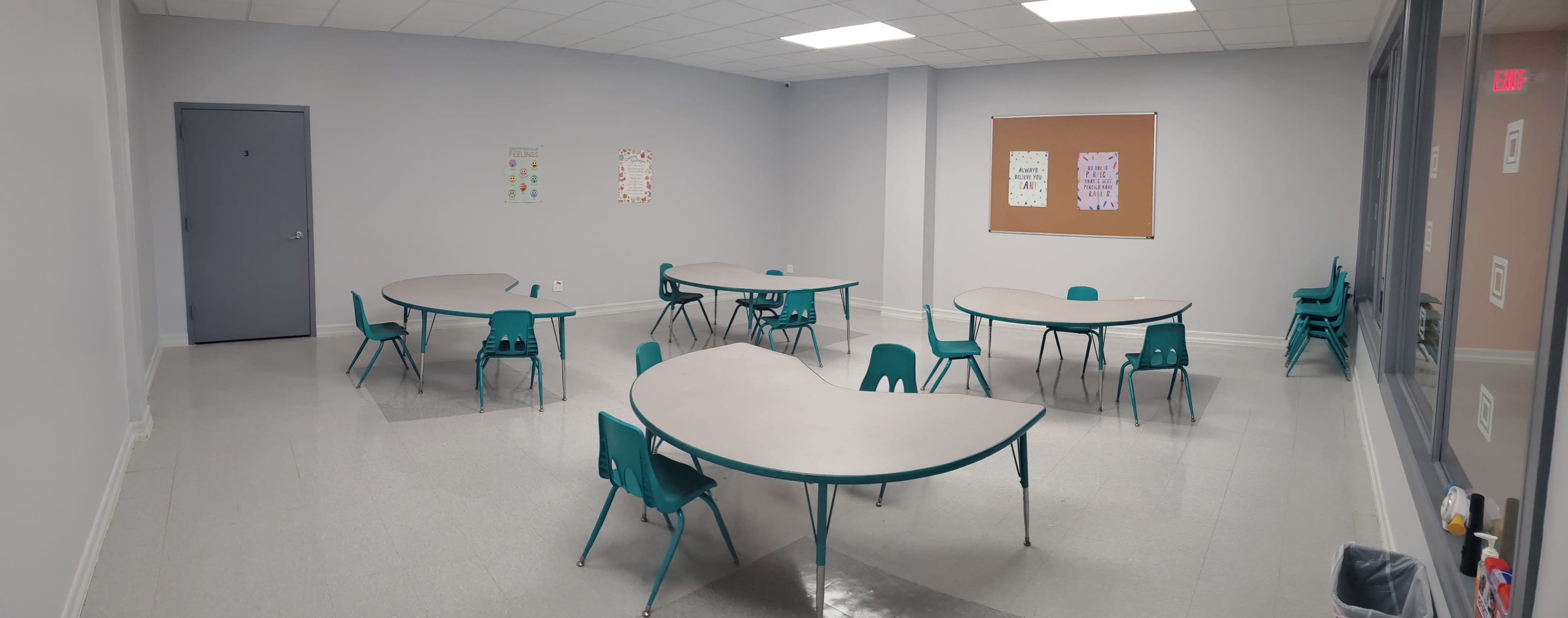 The image shows a classroom with several curved tables and teal chairs arranged neatly in an empty, brightly lit space.