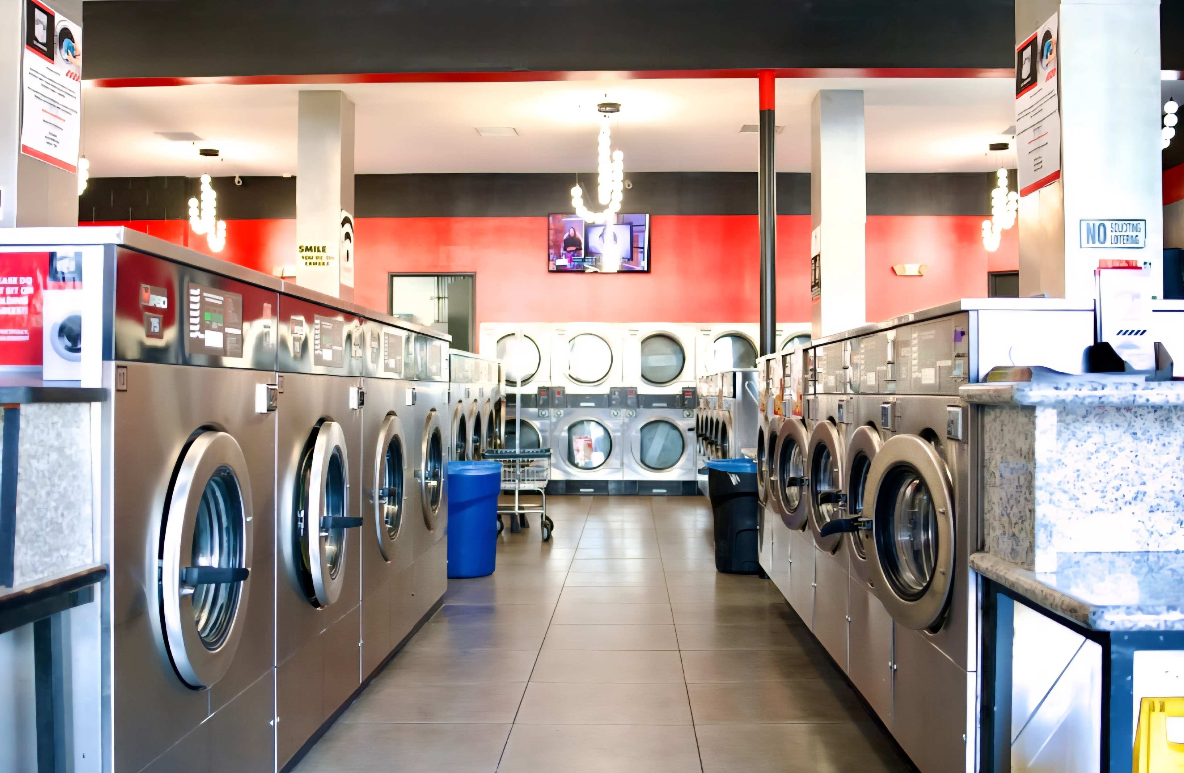 The image shows the interior of a modern laundromat with rows of washing machines, a large window displaying laundry machines, and bright lighting.