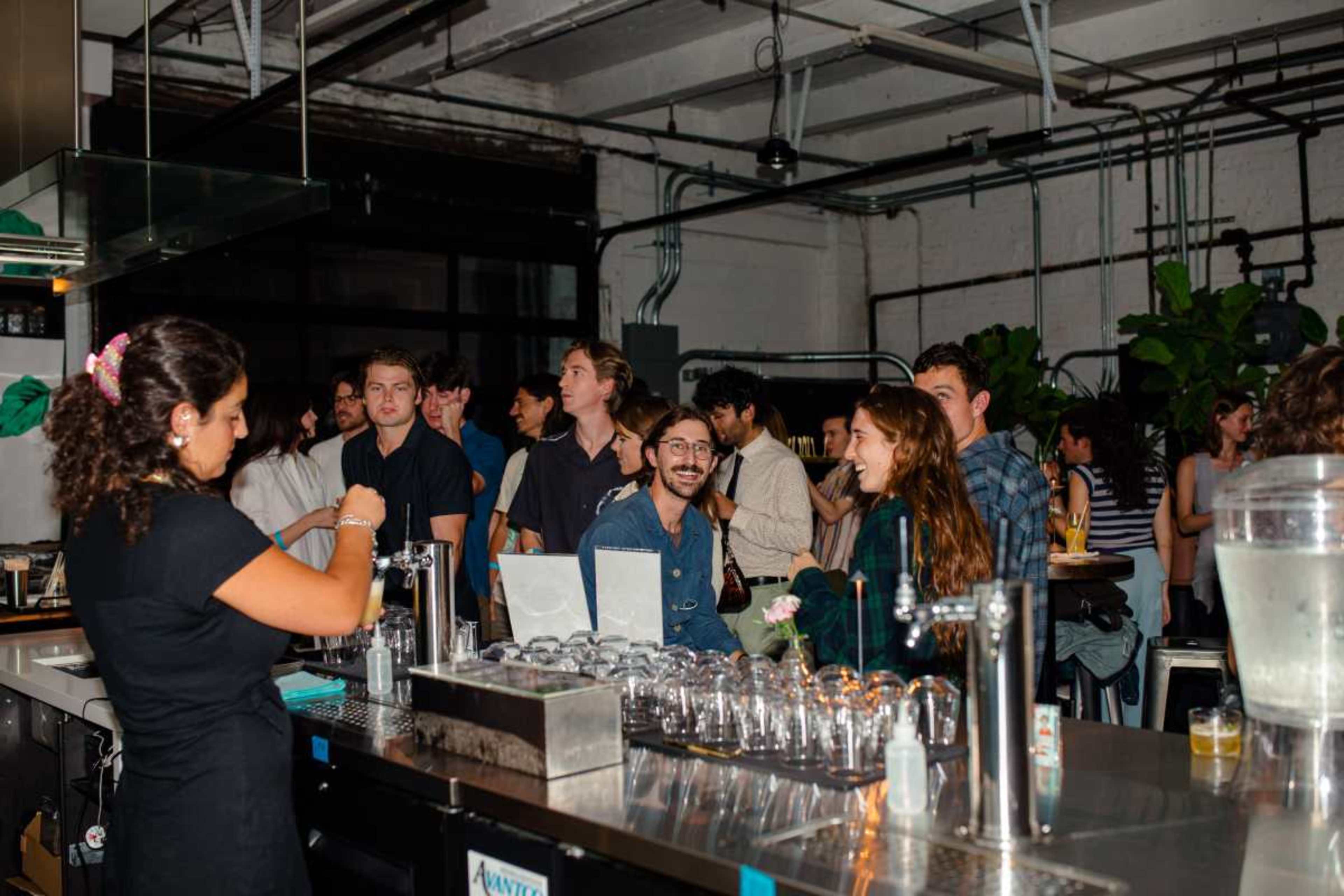 A group of people socializes in a bar setting, with some patrons interacting with a bartender and others engaged in conversation.