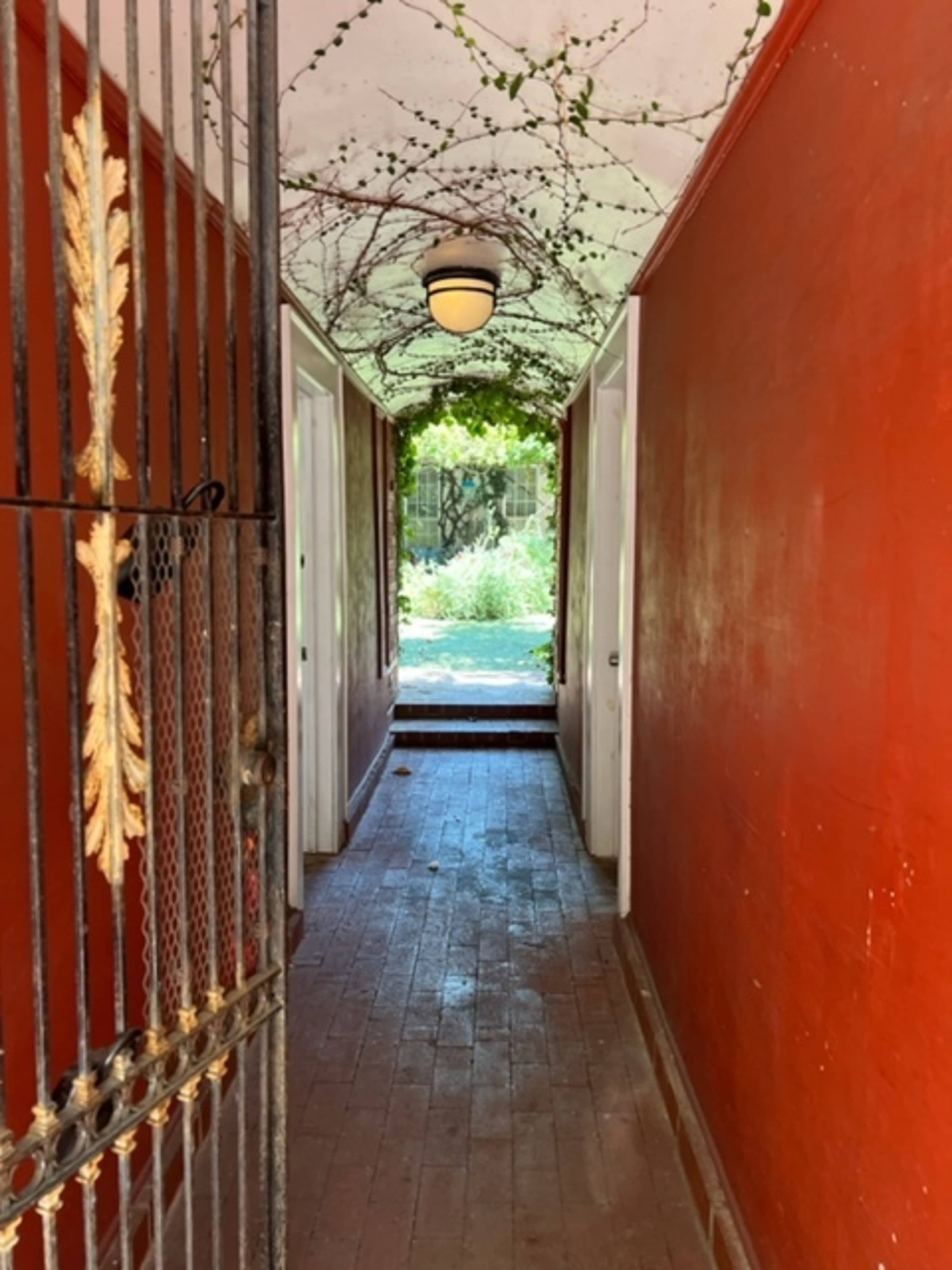 A narrow corridor with red walls and a wrought iron gate leads to a garden visible through an archway.