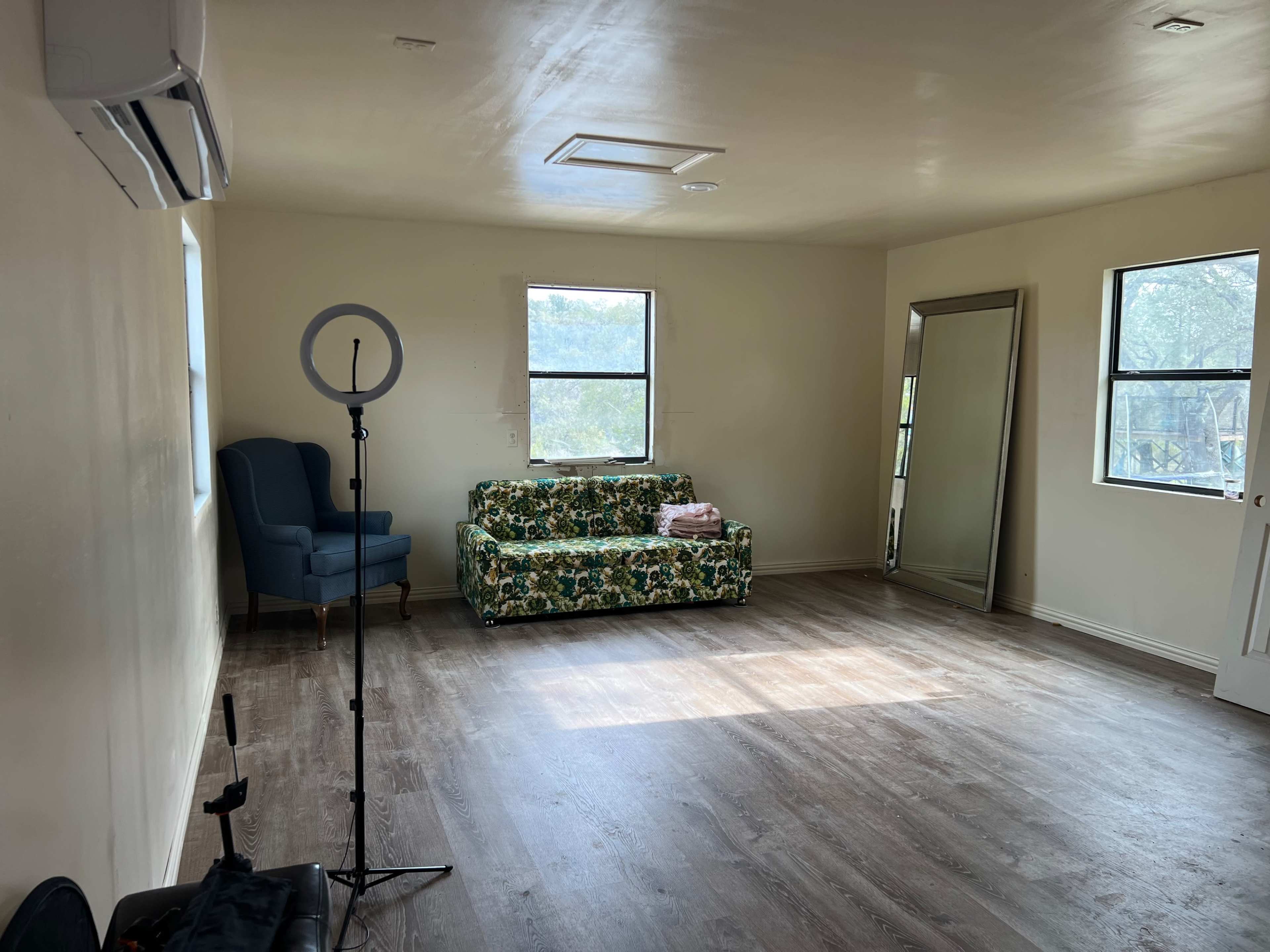 The image shows a vacant room with a floral-patterned sofa, a tall mirror, and a ring light setup beside an armchair.