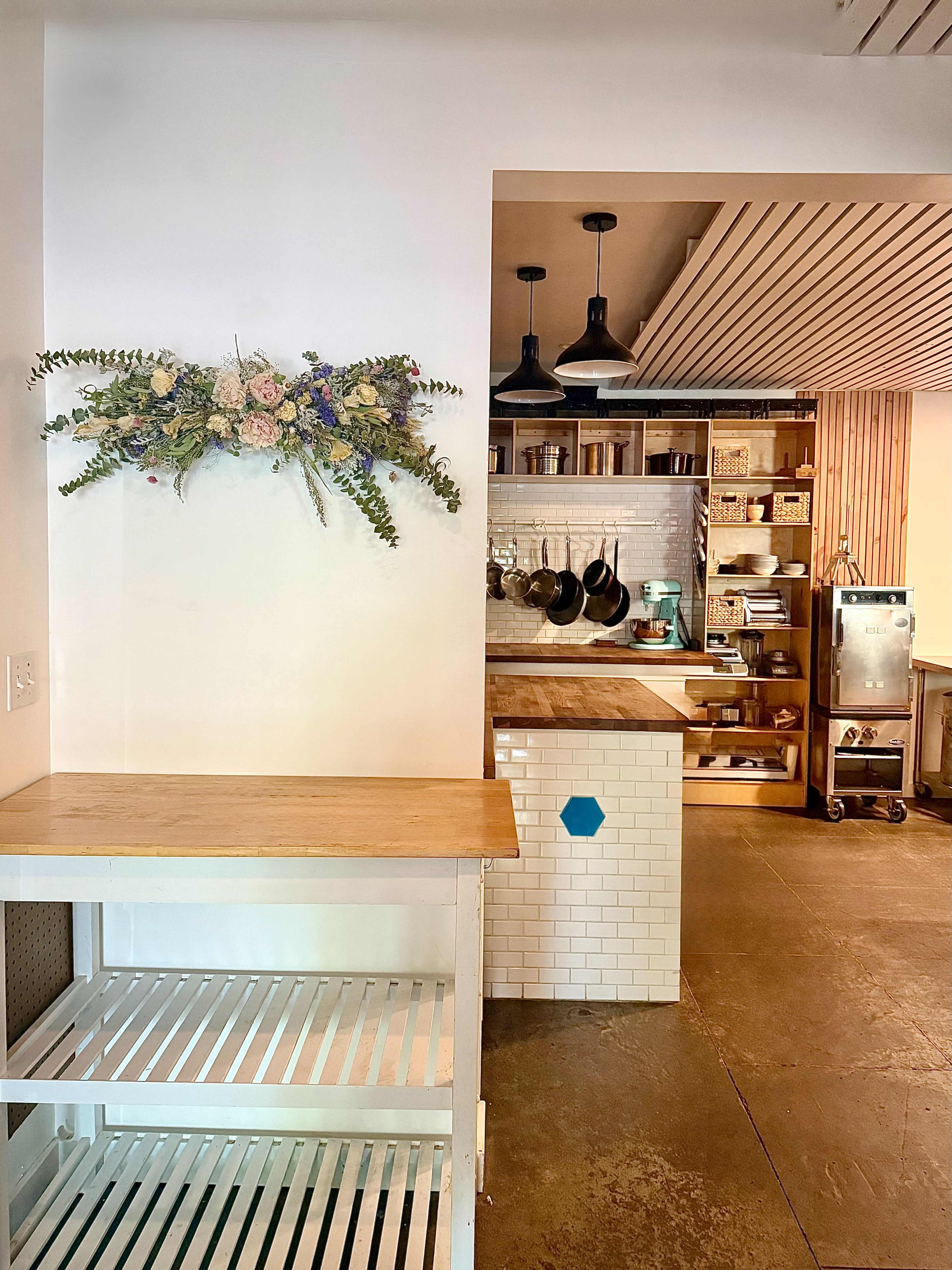 The image shows a stylish kitchen with wooden elements and a wall-mounted floral arrangement, featuring an open layout that connects to a dining area.