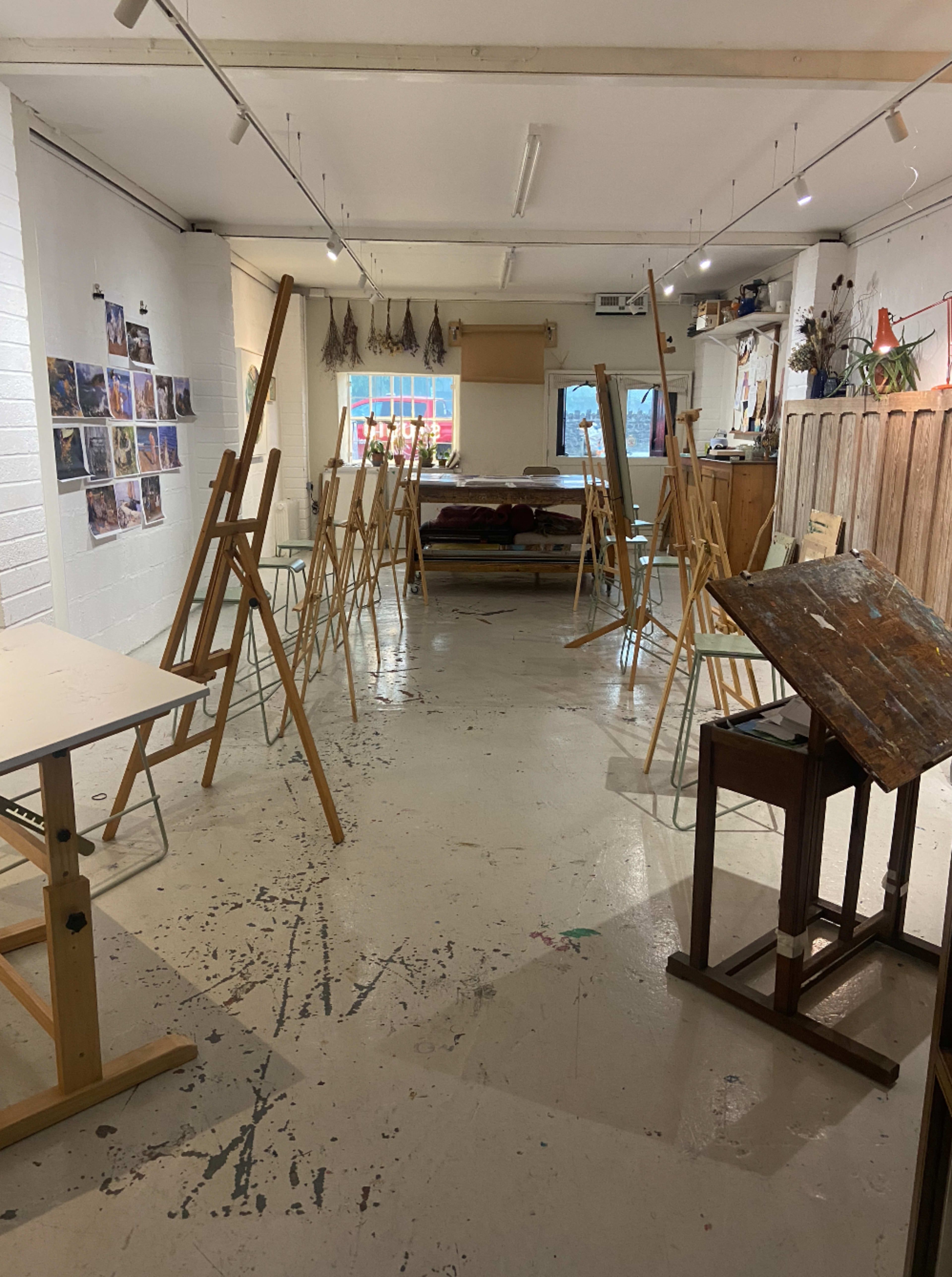 The image shows an art studio with several wooden easels arranged throughout the space, along with a wooden table and various artworks displayed on the walls.