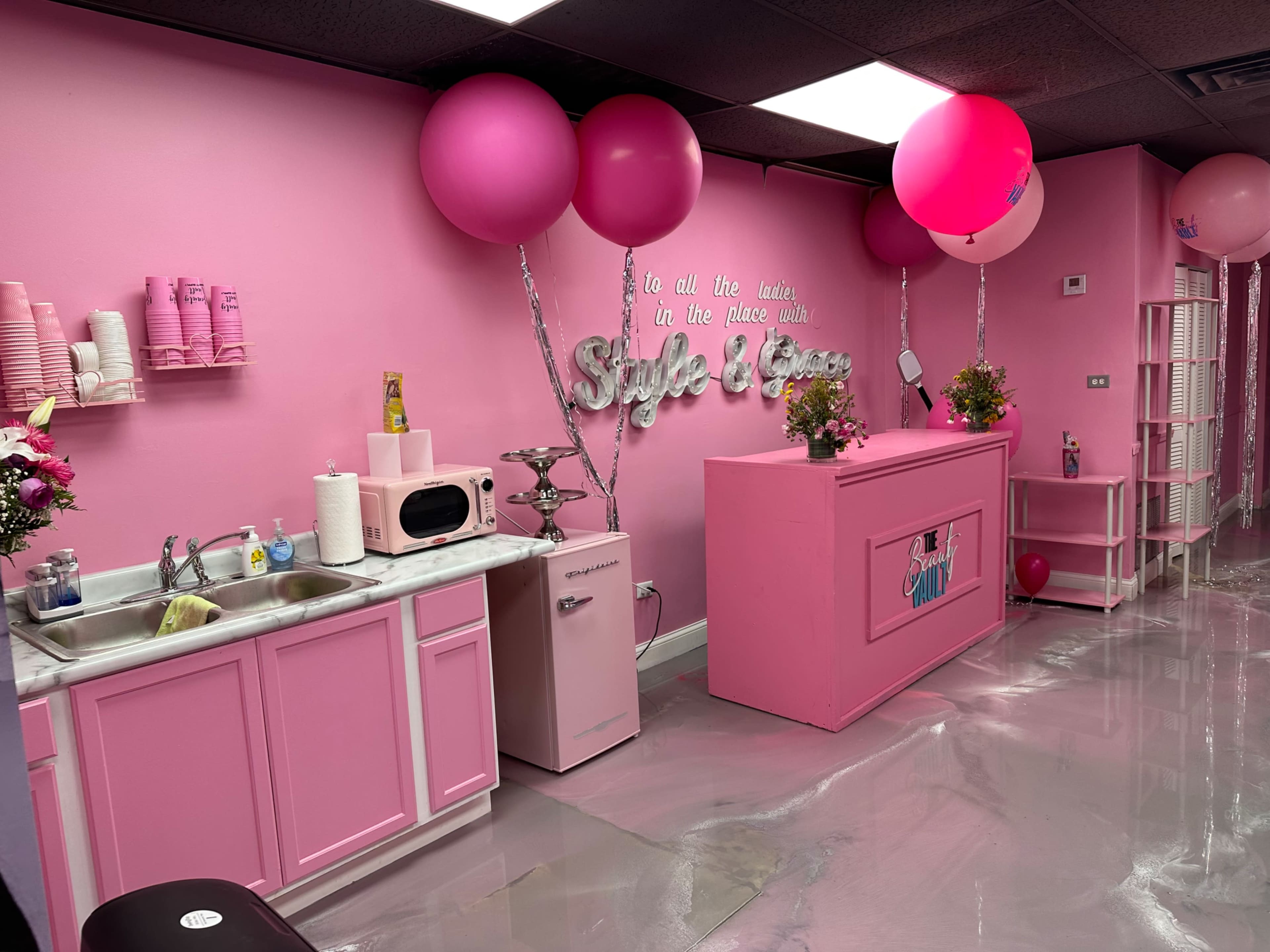 A brightly colored pink interior of a beauty salon with a kitchenette area, decorated with balloons and flowers.