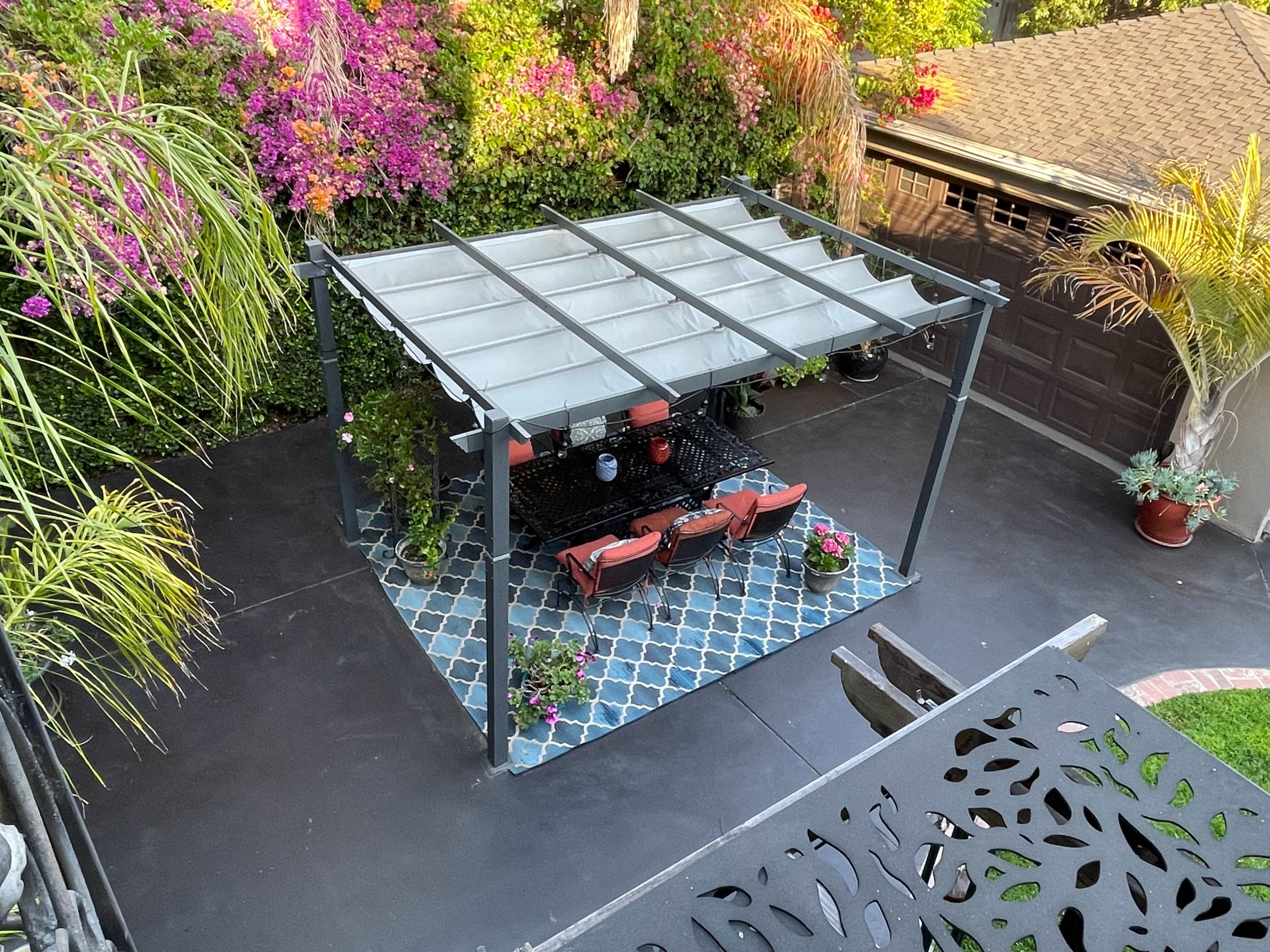 The image shows a patio with a covered seating area surrounded by colorful plants and flowers, with a patterned floor and a garage in the background.