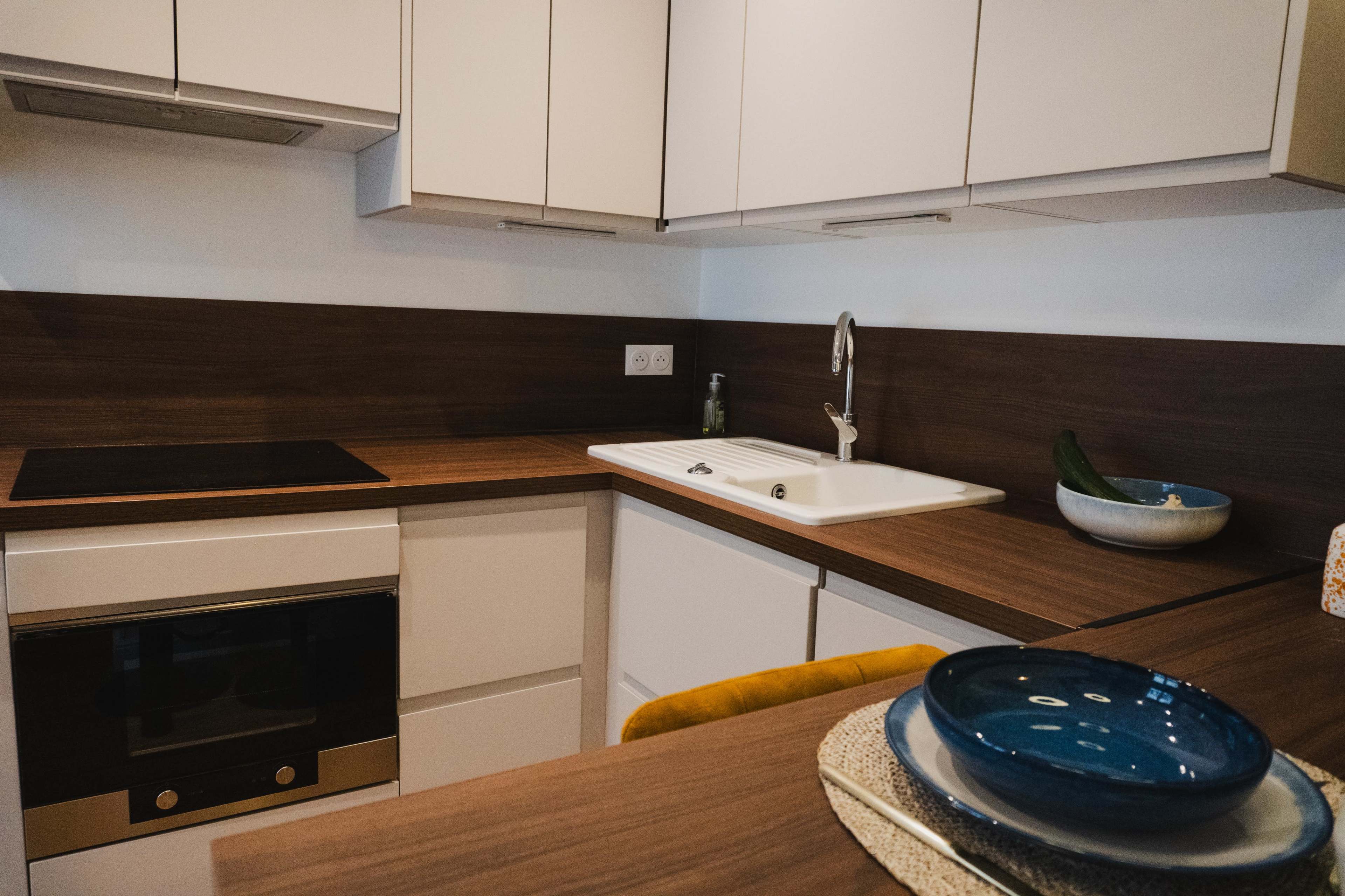 The image shows a modern kitchen with a countertop, sink, and built-in oven, featuring sleek cabinetry and dishware arranged on the surface.