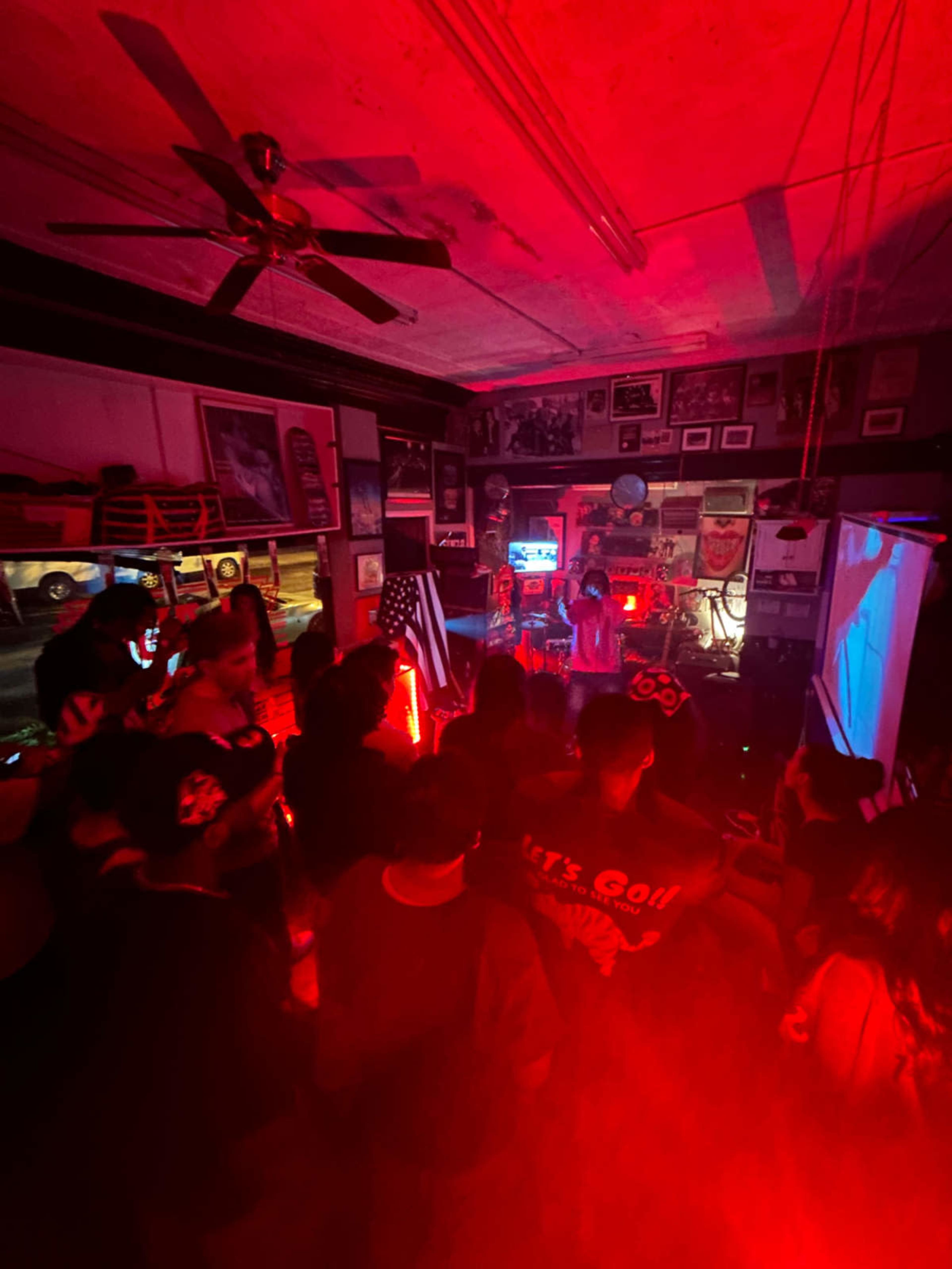 A crowd gathers in a dimly lit room adorned with posters and memorabilia while a performer stands on a small stage under red lights.