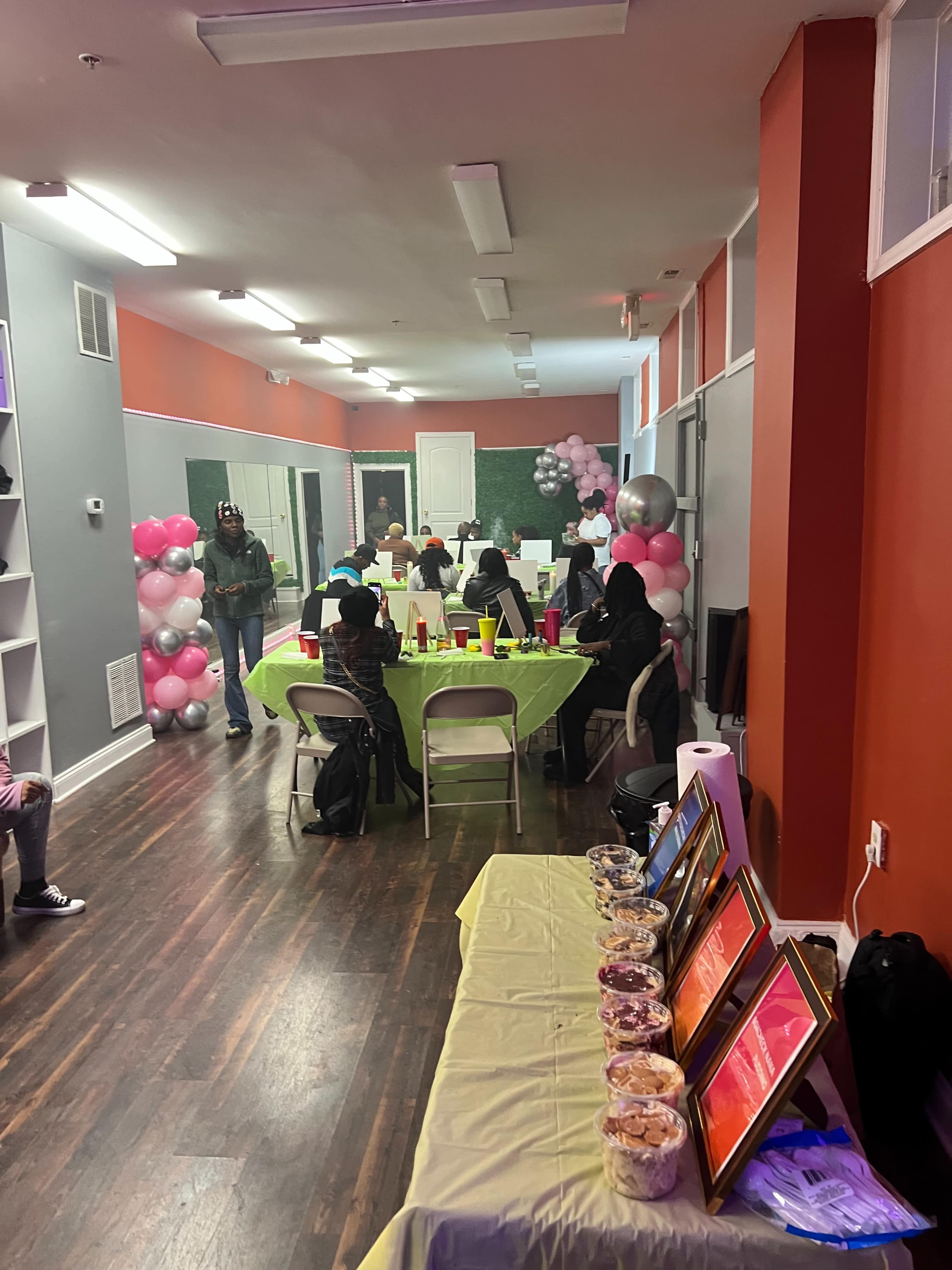 A decorated event space with tables covered in bright green cloth, a buffet of snacks, and balloons along the walls, while guests sit around enjoying the gathering.