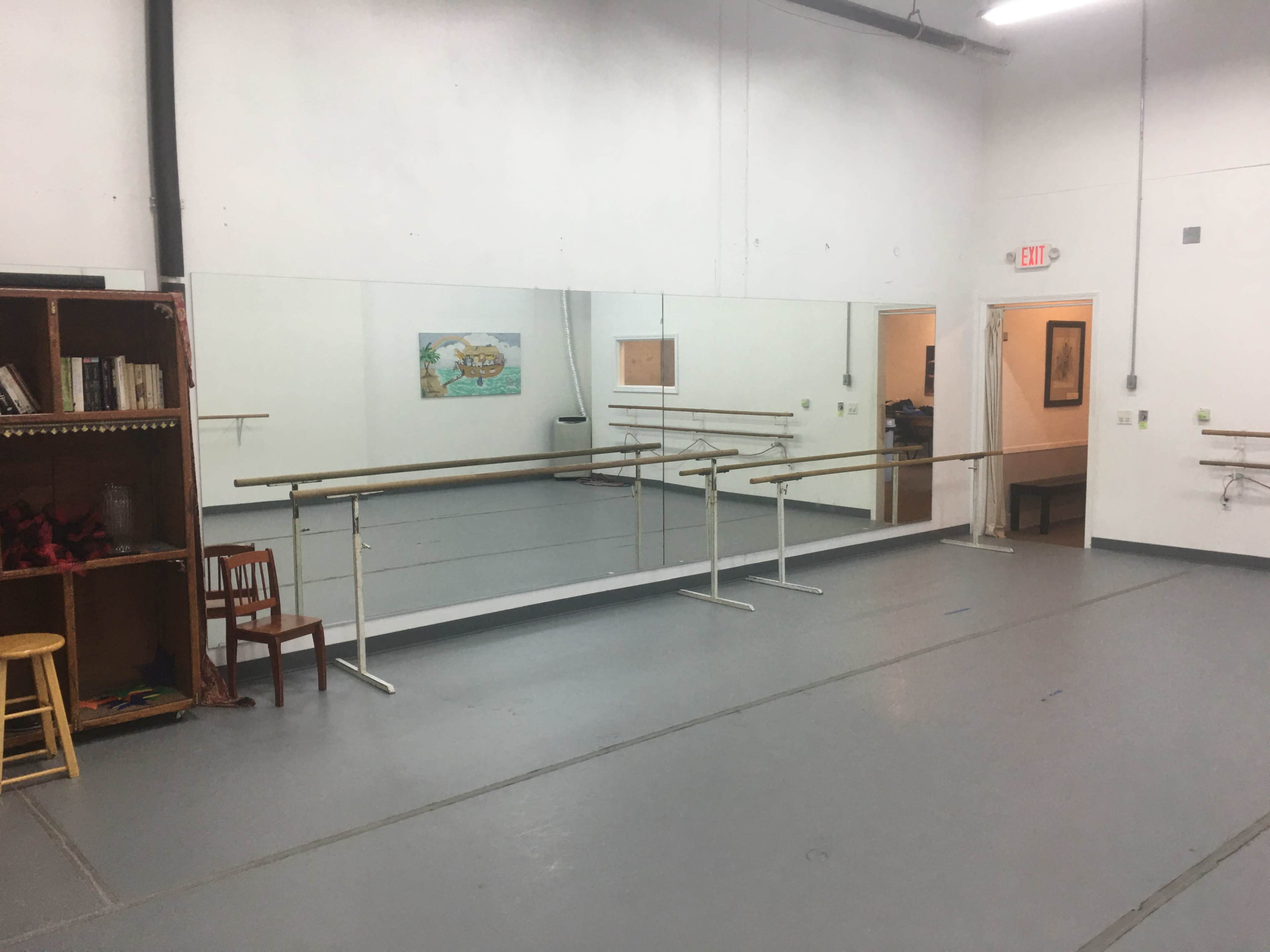 A dance studio with mirrored walls, ballet barres, and a wooden chair near a bookshelf.