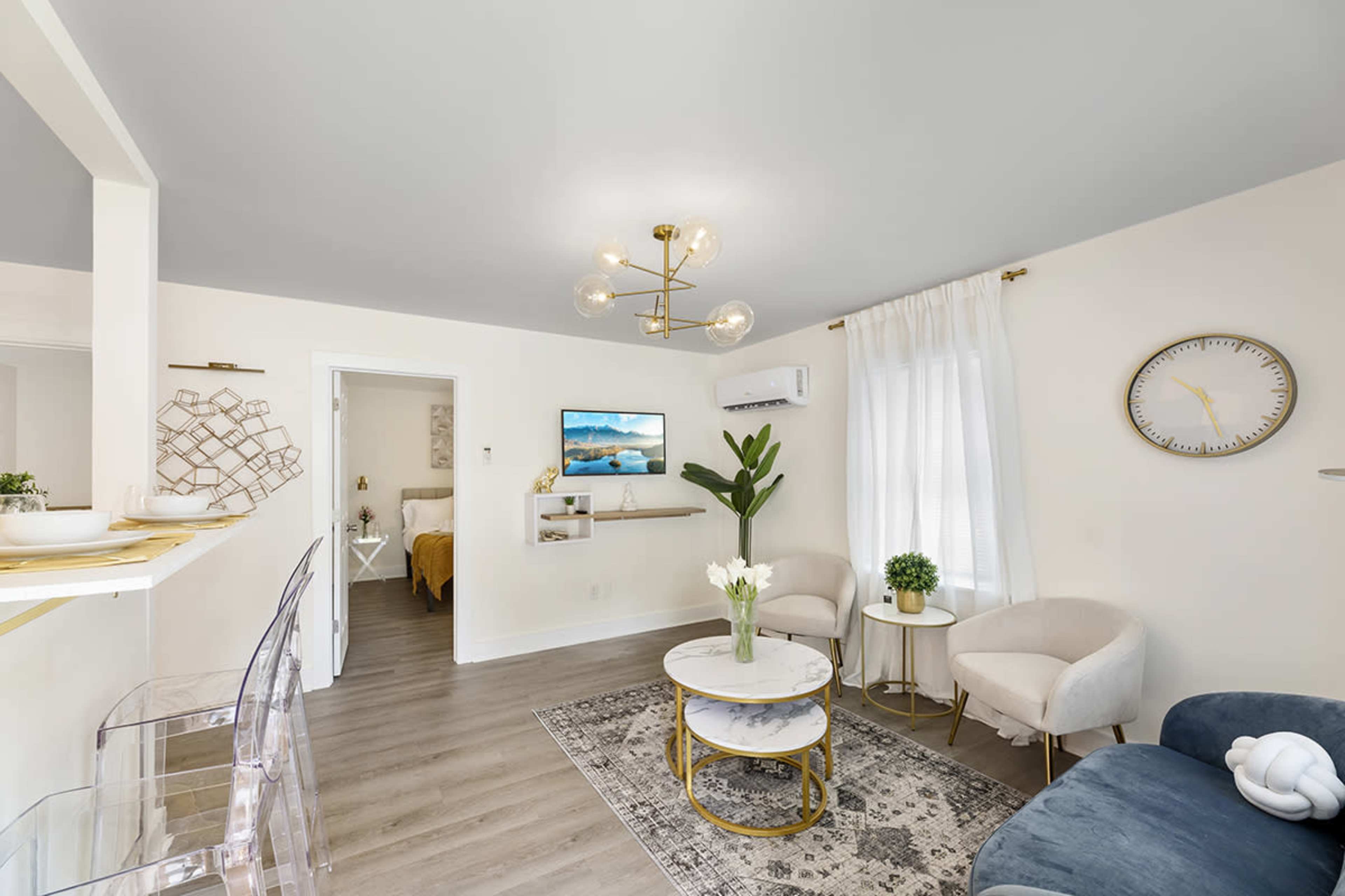 A modern living room with a coffee table, two accent chairs, a wall-mounted television, and a view of a separate bedroom through an open doorway.