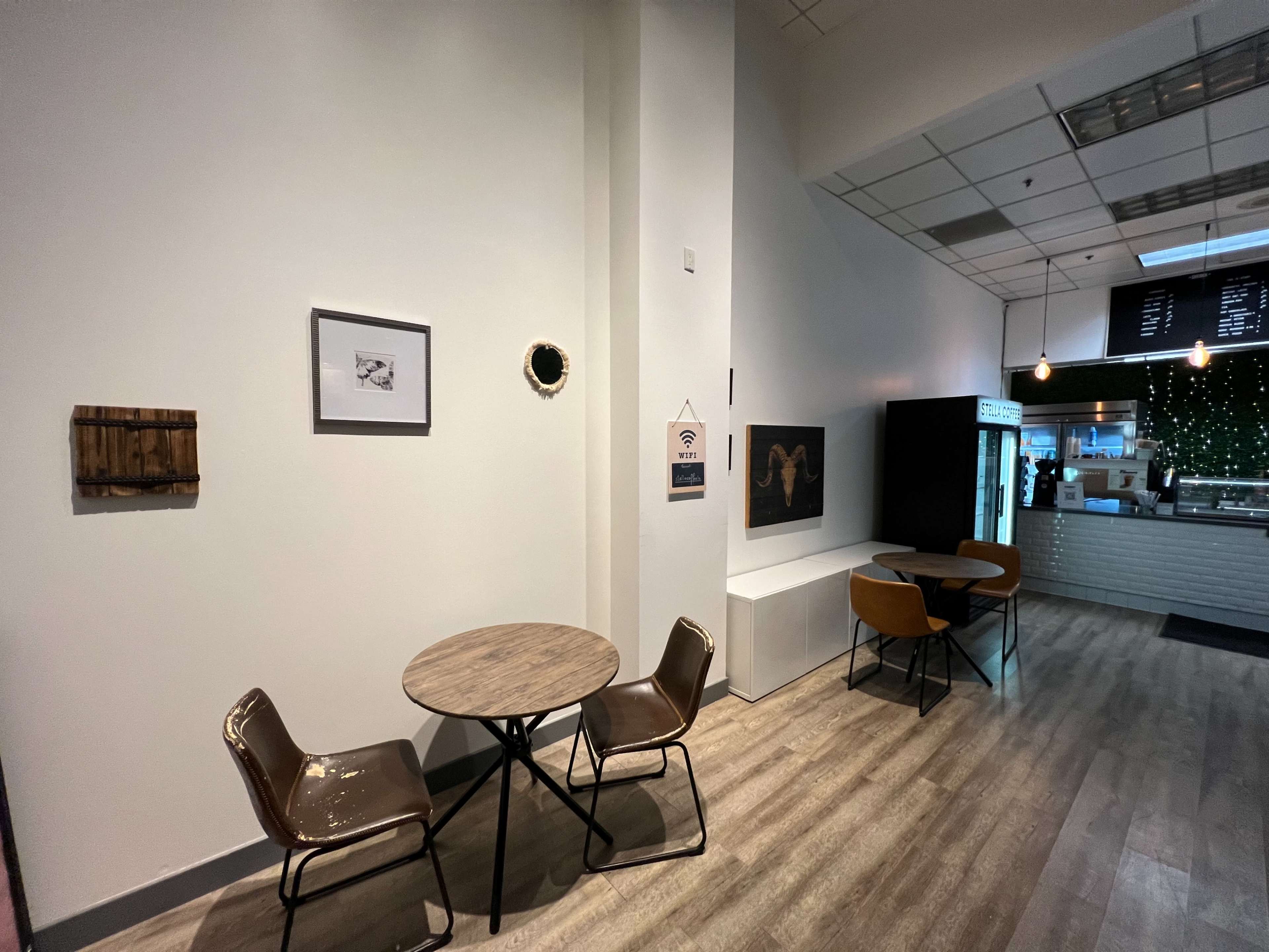 The image shows a small cafe area with two tables and chairs against a white wall, featuring a few pieces of wall art and a visible counter in the background.