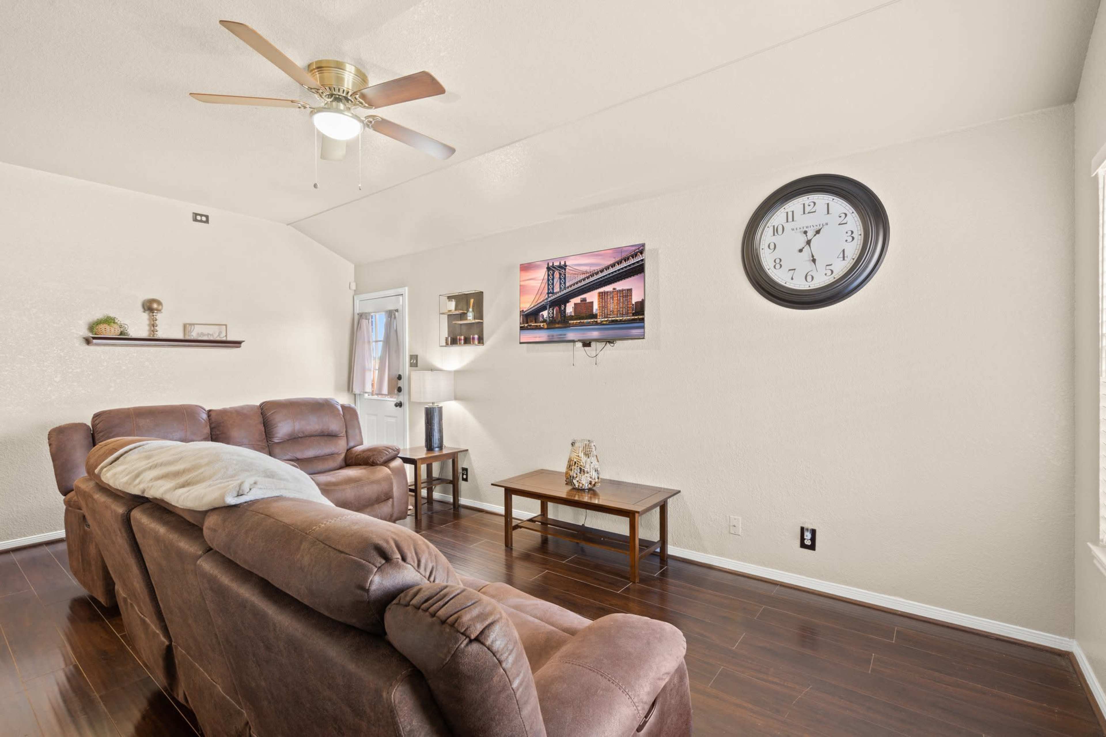 A living room features a brown sectional sofa, a wall-mounted television, a clock, and a small table on a hardwood floor.