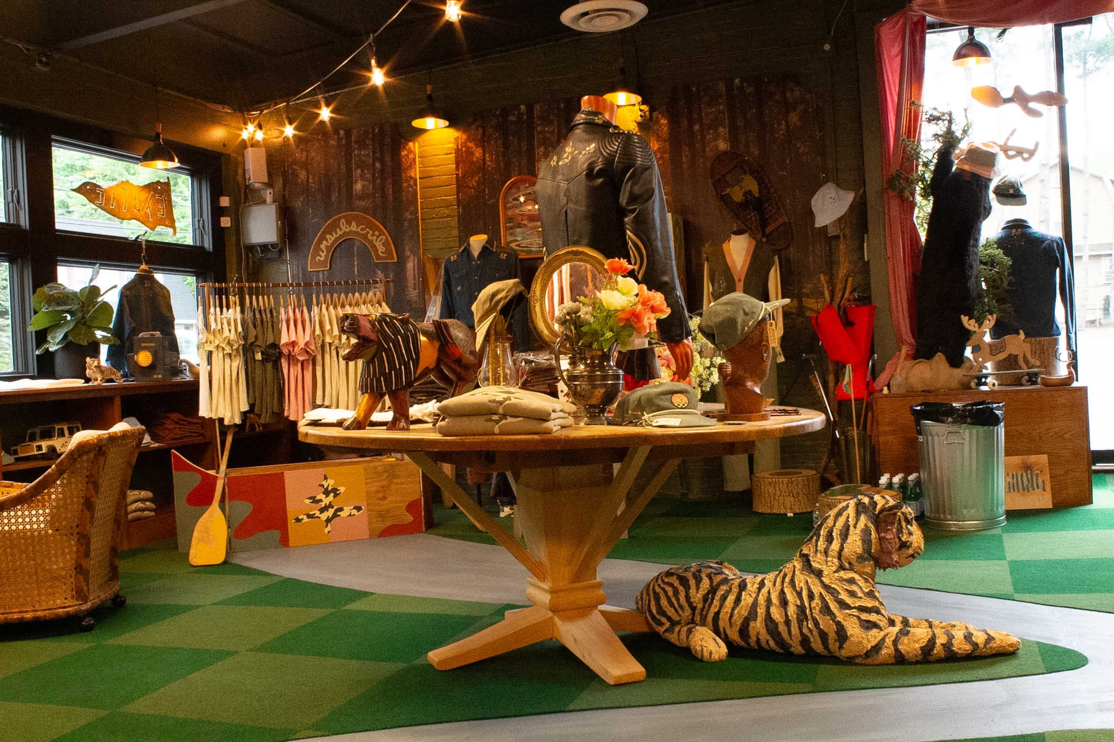 The image shows a boutique store interior featuring clothing displays, a wooden table with hats and accessories, and a striped plush tiger on the floor amidst a rustic décor.