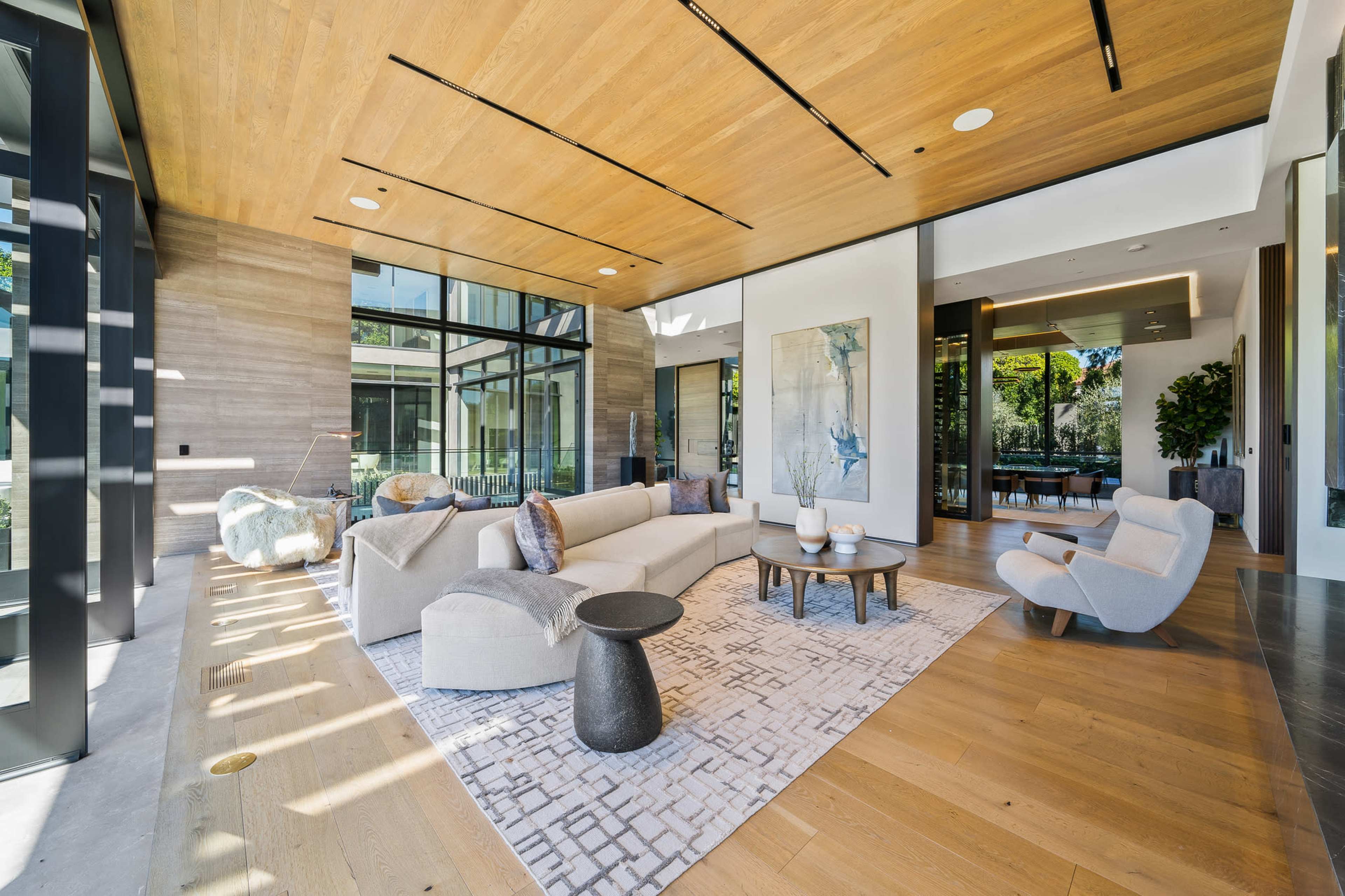 The image shows a modern living room with large windows, light wood flooring, and contemporary furniture arranged around a central rug.