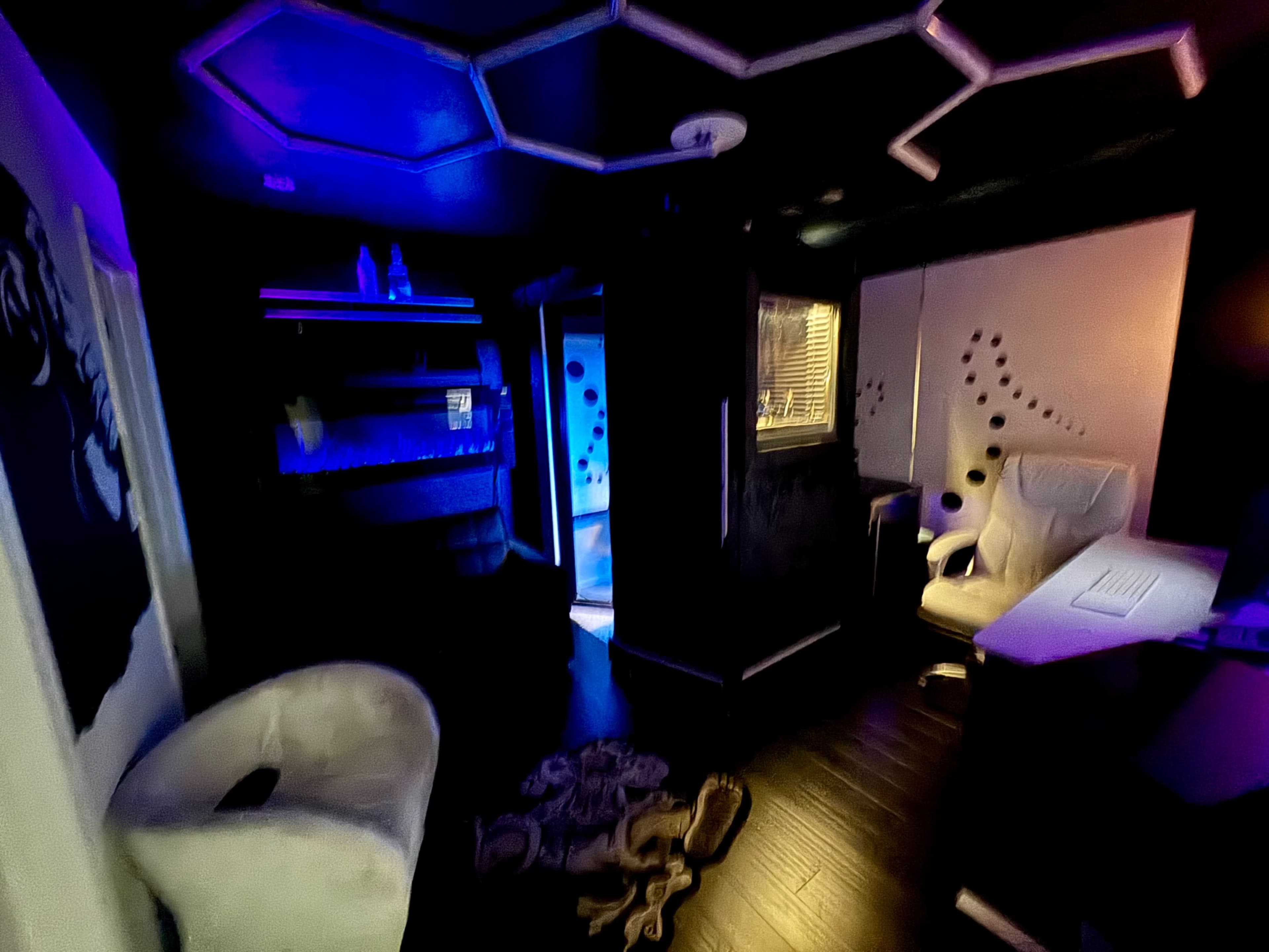 The image shows a dimly lit room with black walls, featuring modern furniture, including a white chair and a desk, alongside blue and purple lighting effects.