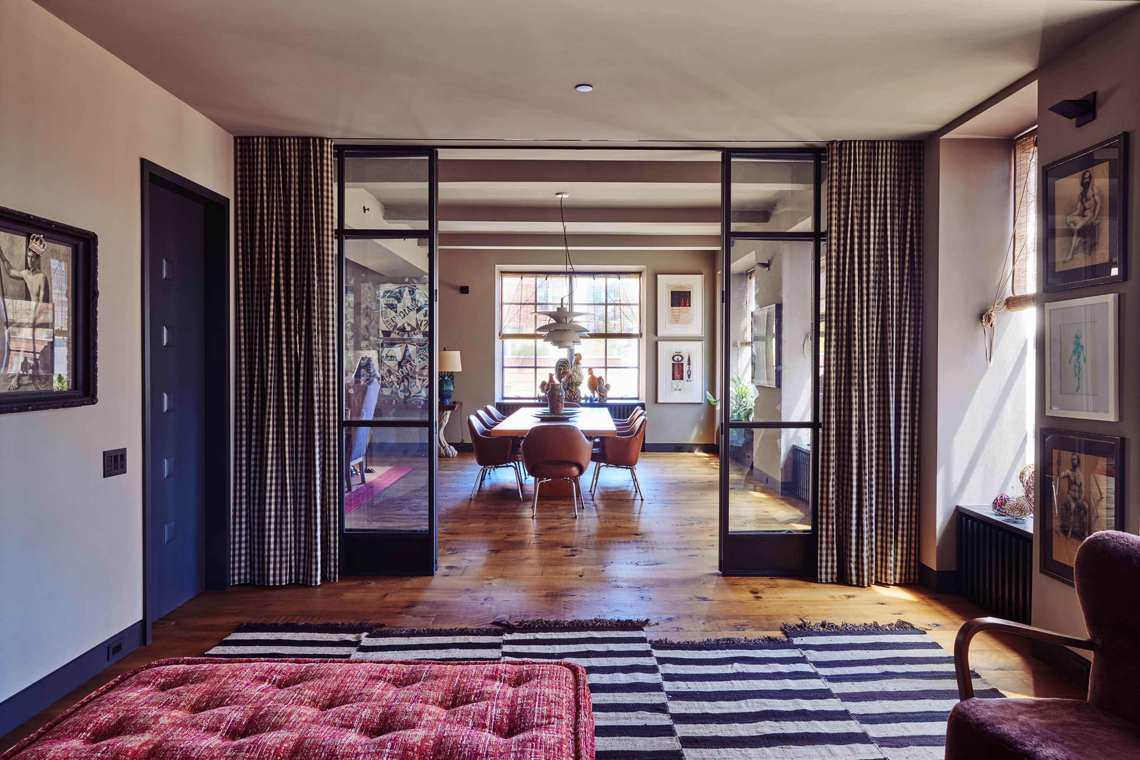 A modern living room features a view into a dining area, separated by glass doors, with a large dining table and stylish furnishings.