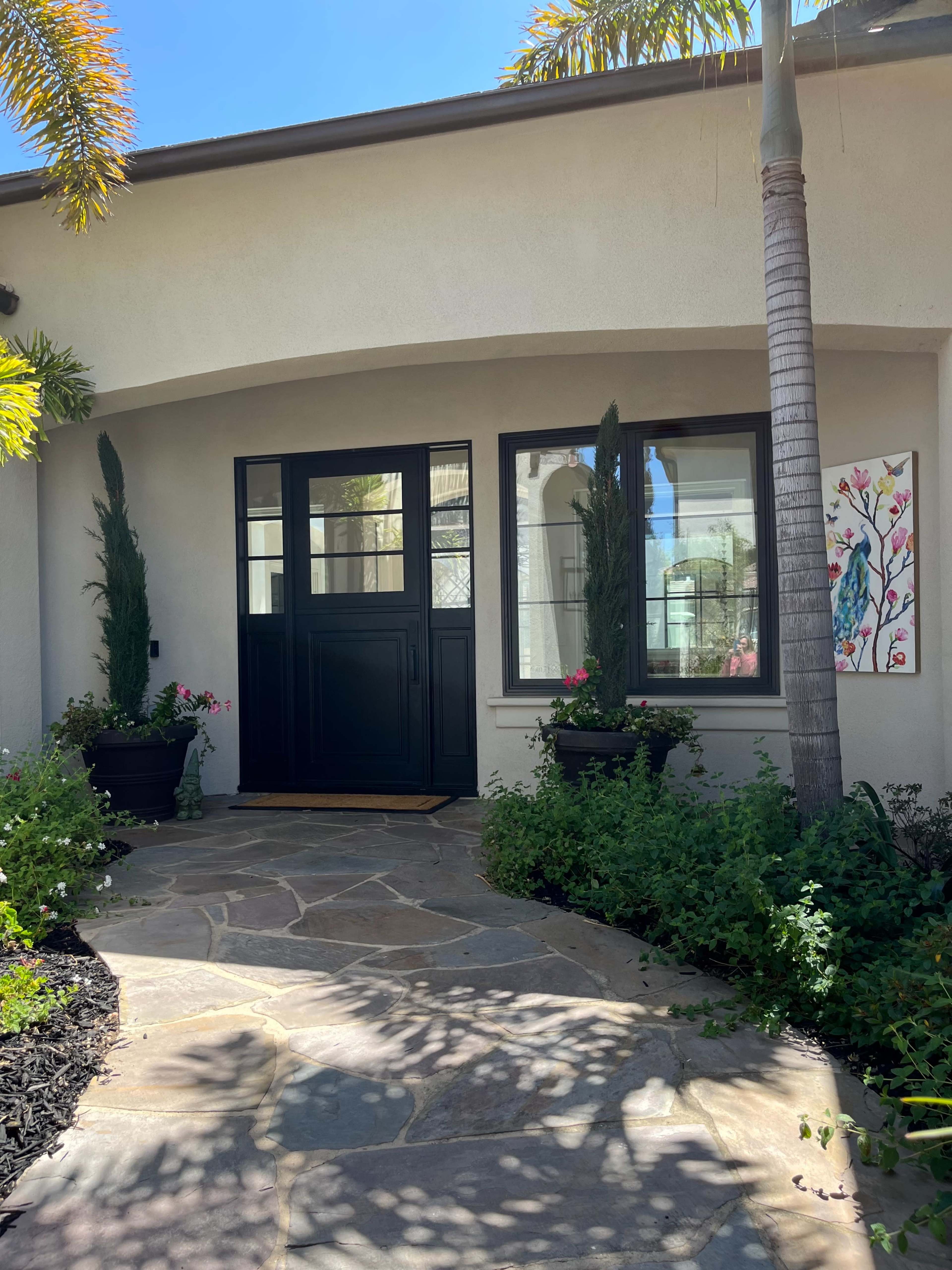 A pathway made of stone leads to a black front door of a house, flanked by potted plants and a colorful artwork on the side wall.