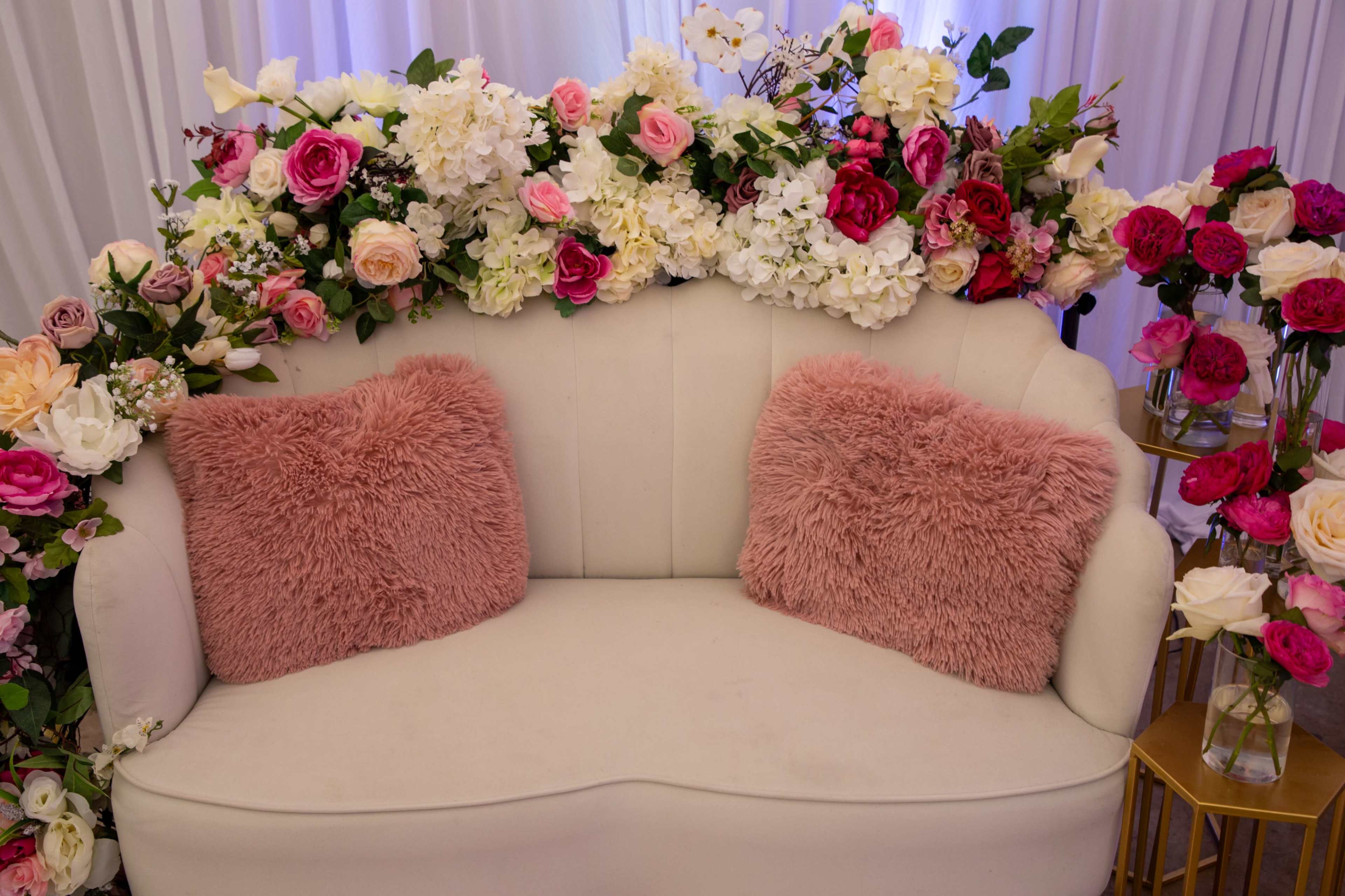 A cream-colored loveseat is adorned with plush pink pillows and surrounded by a variety of colorful artificial flowers.