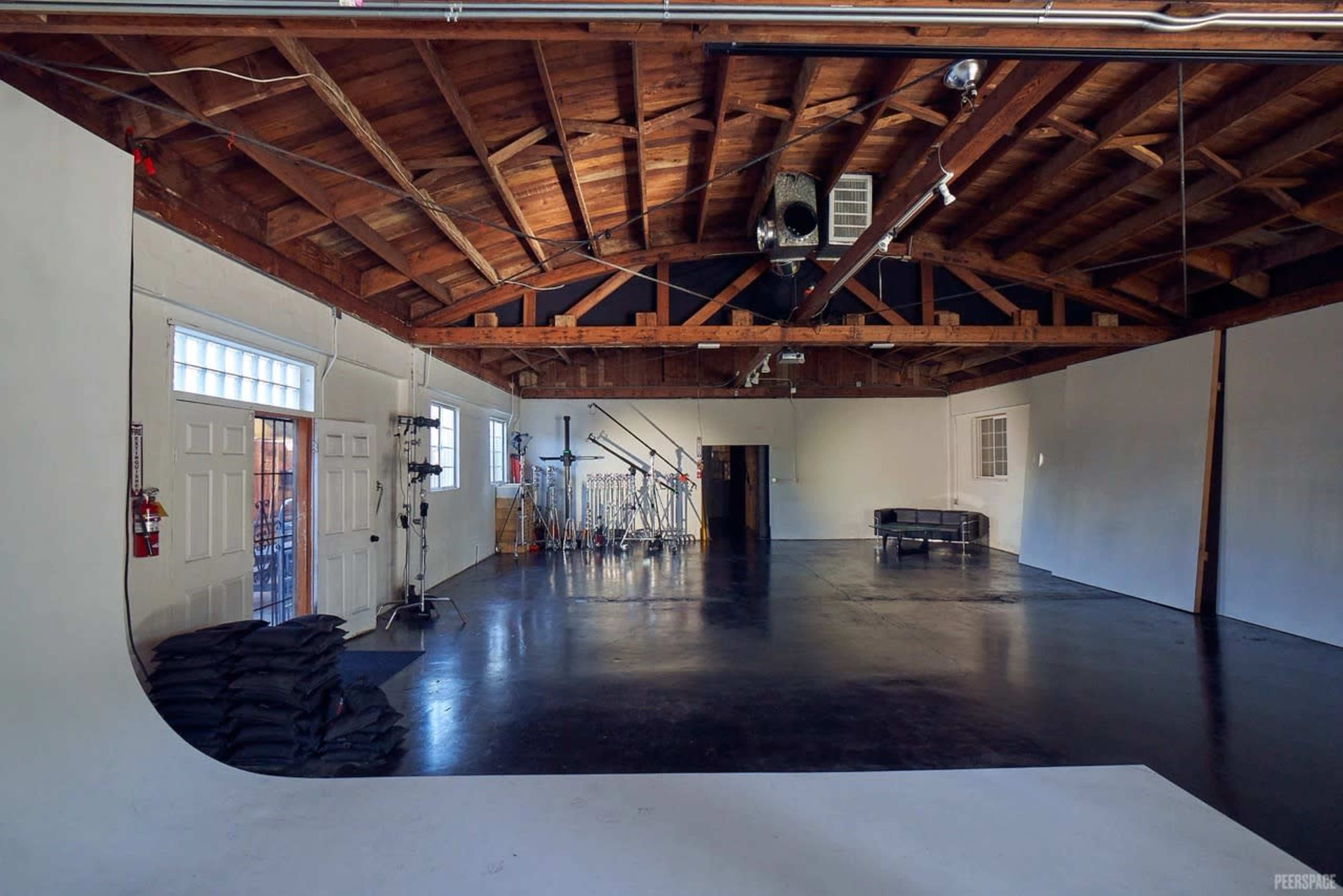 The image shows a spacious, empty studio with a wooden ceiling, a black floor, and various equipment set against the back wall.