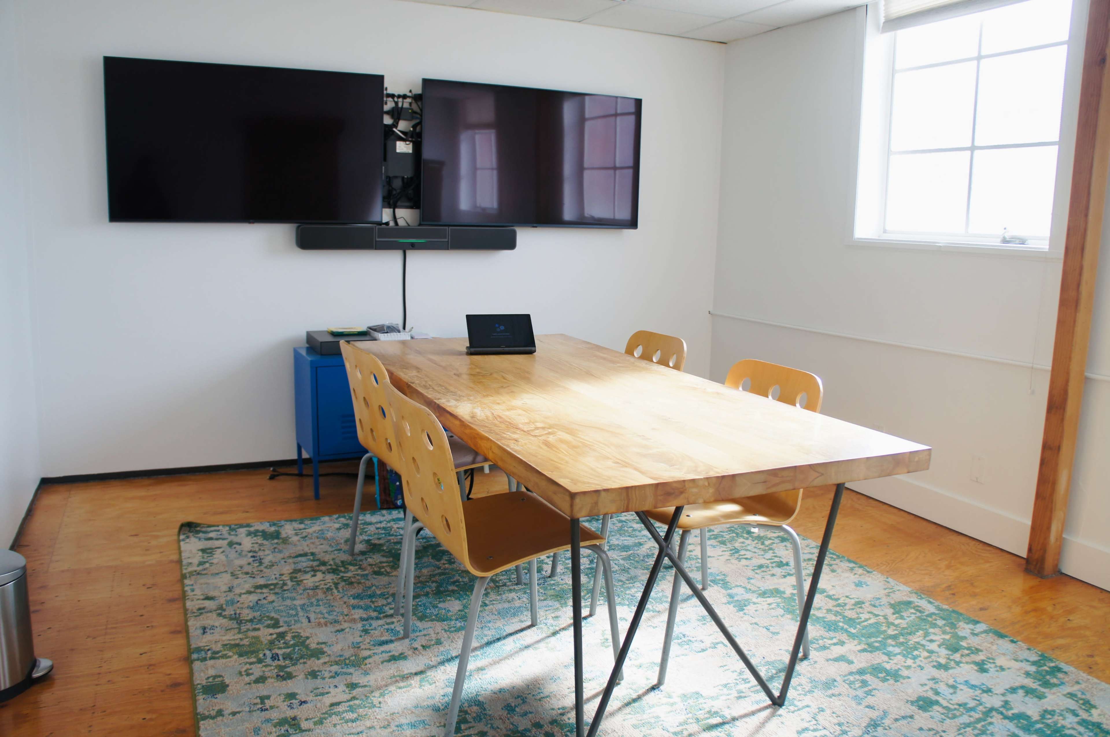 A conference room features a large wooden table surrounded by four orange chairs, with two wall-mounted televisions and a blue cabinet in the background.