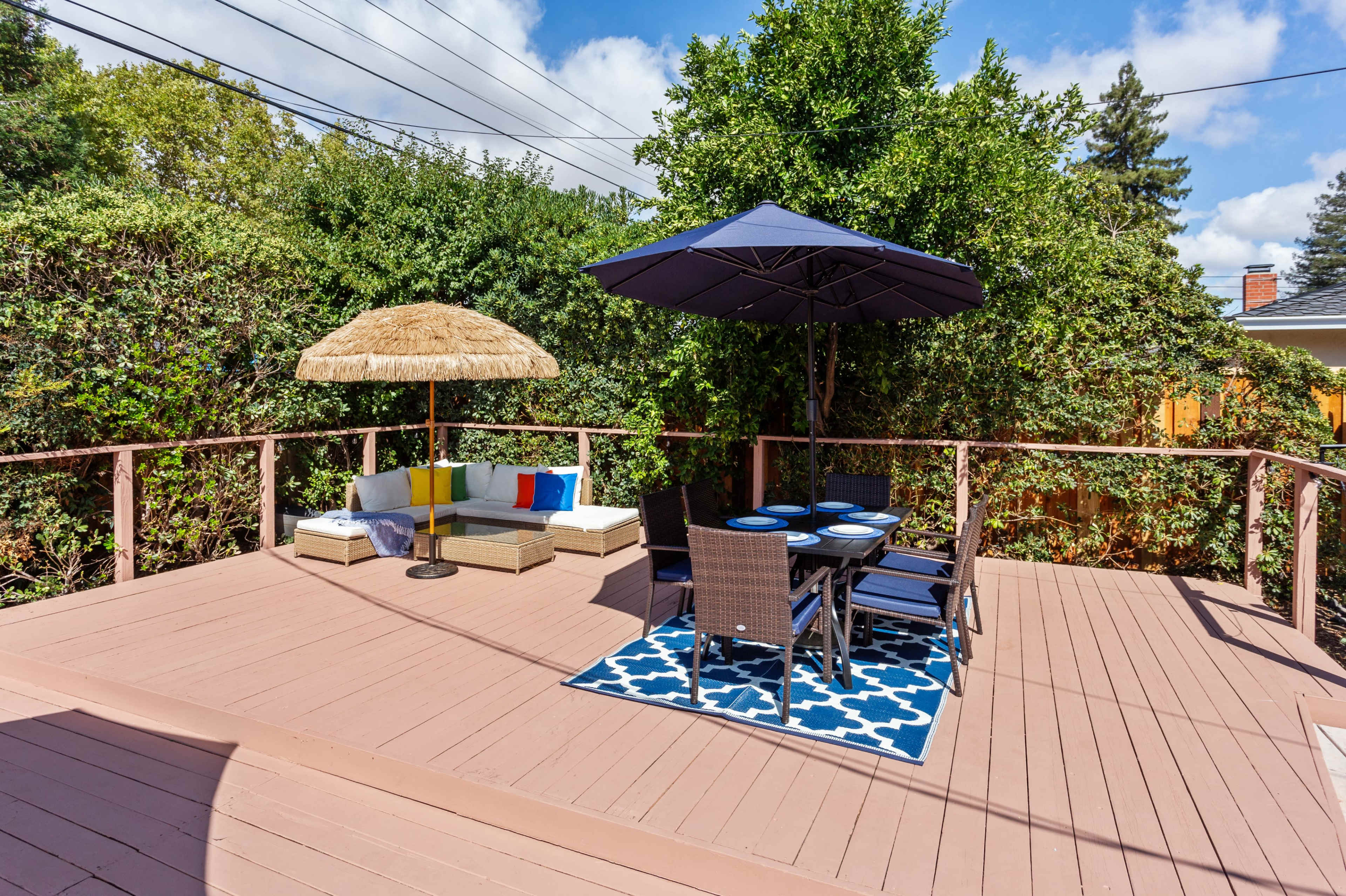 A wooden deck features a seating area with a sofa and a table set under an umbrella and a thatched roof structure.