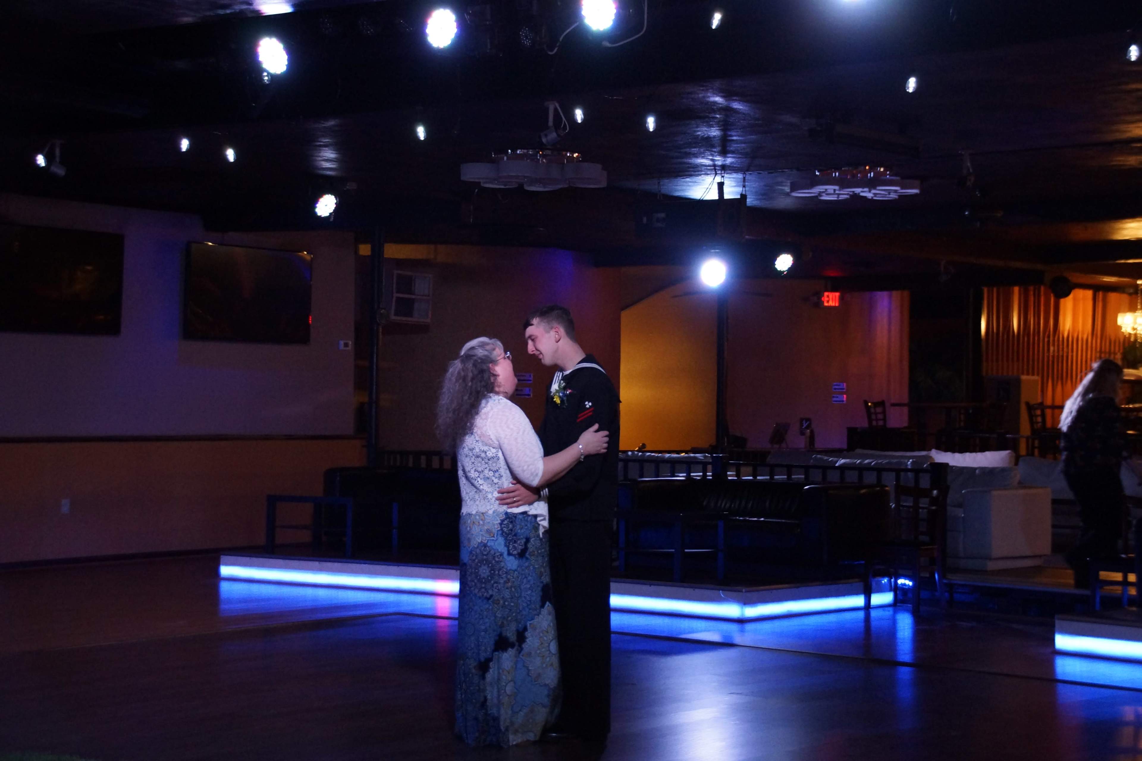 A couple dances closely on a dimly lit dance floor, surrounded by ambient lighting and empty tables in the background.