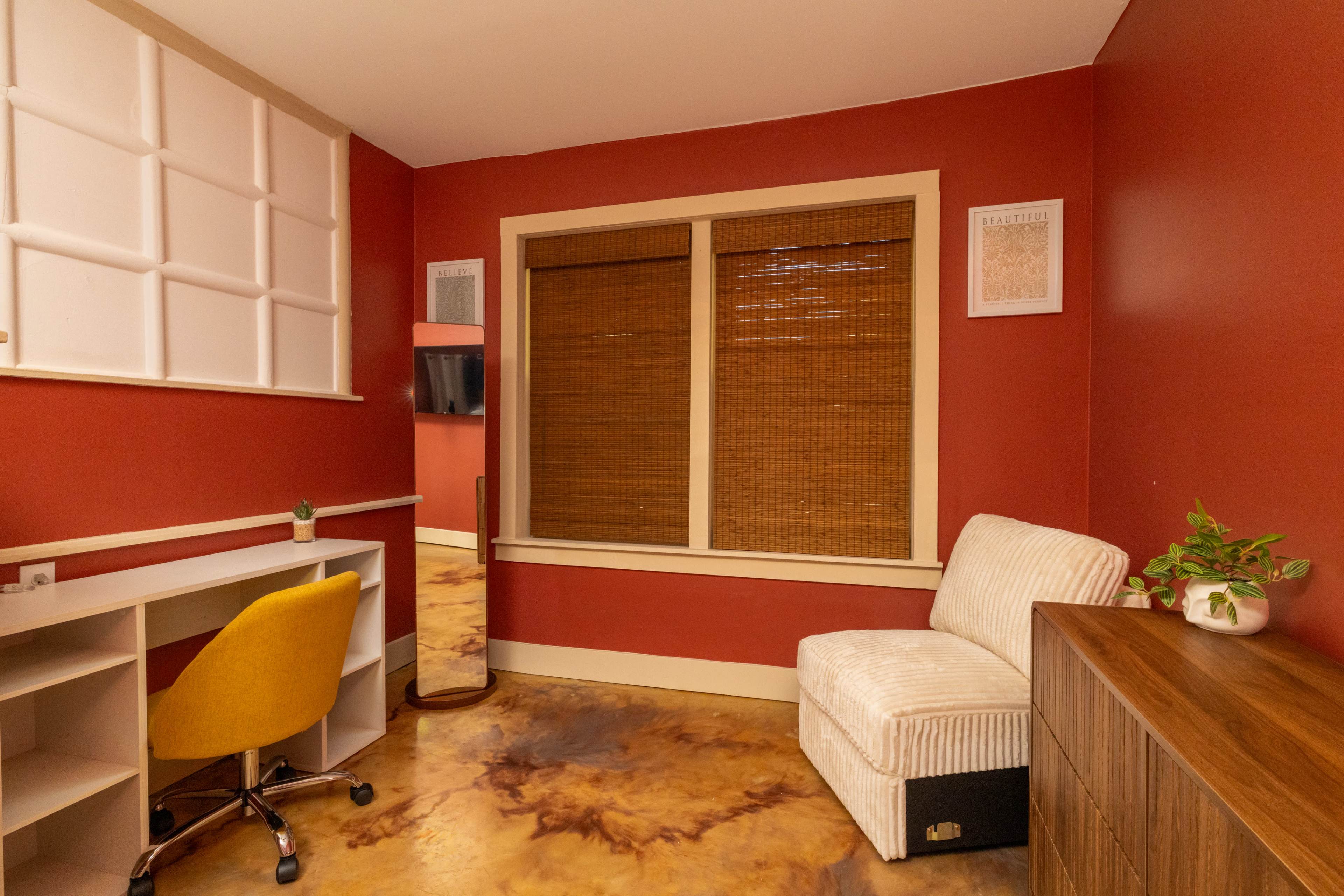 The image shows a small room featuring a desk with a yellow chair, a white couch, and a window with wooden blinds, all set against red walls.