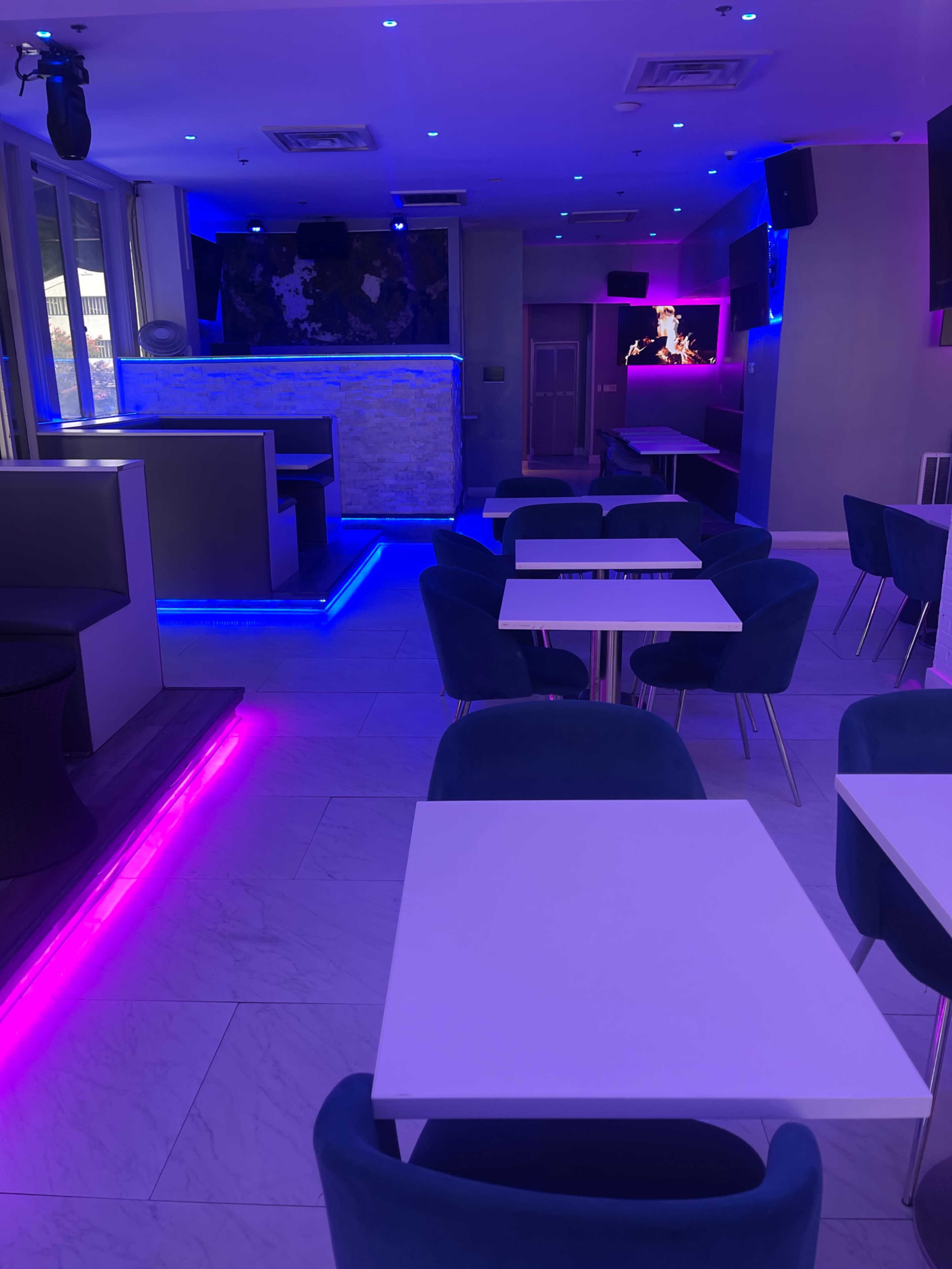 A modern, well-lit lounge with blue and pink ambient lighting, featuring several empty tables and a bar area in the background.