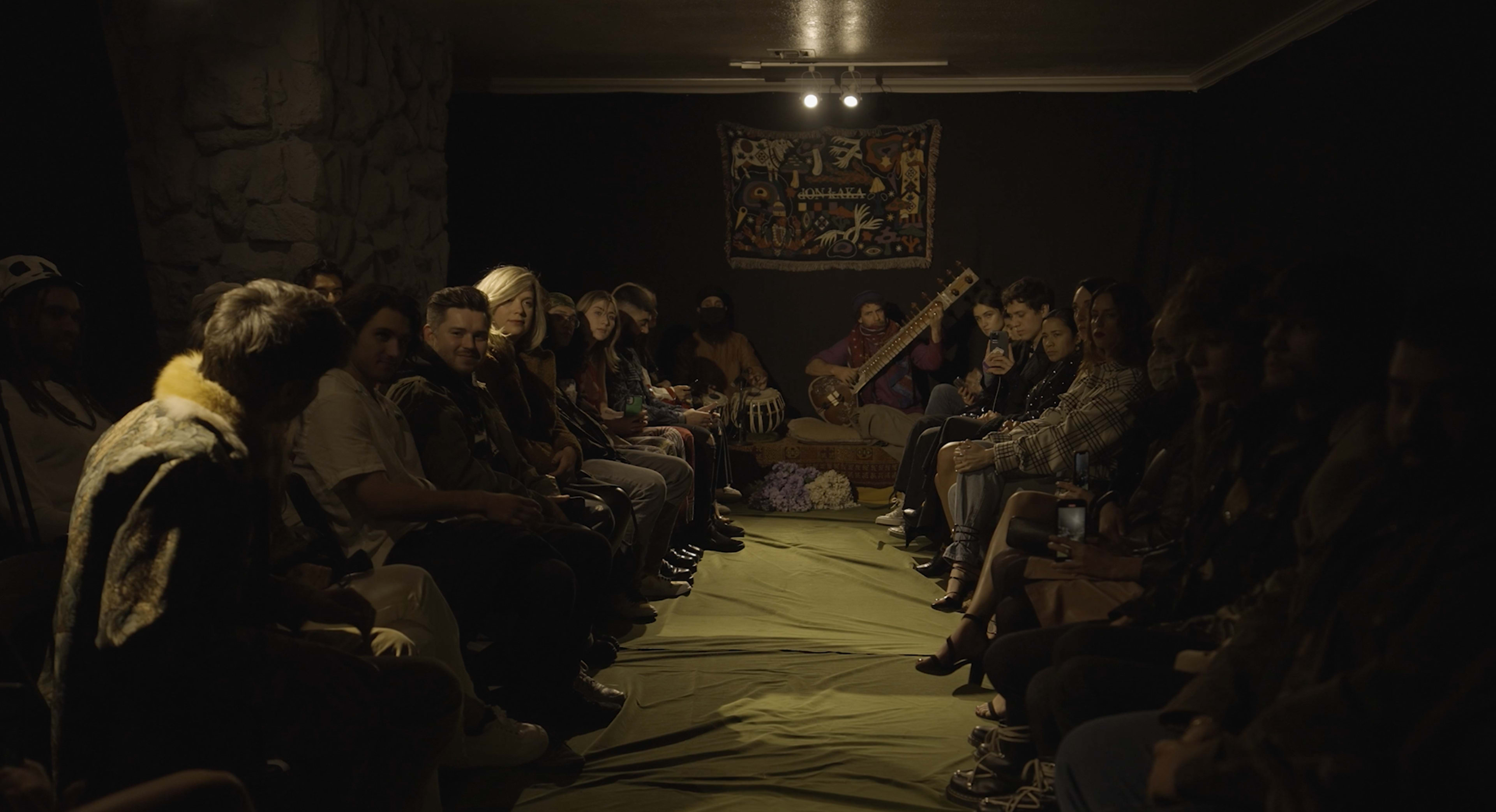 A group of people sits in two rows on a green mat in a dimly lit room, while a musician plays an instrument at the front.