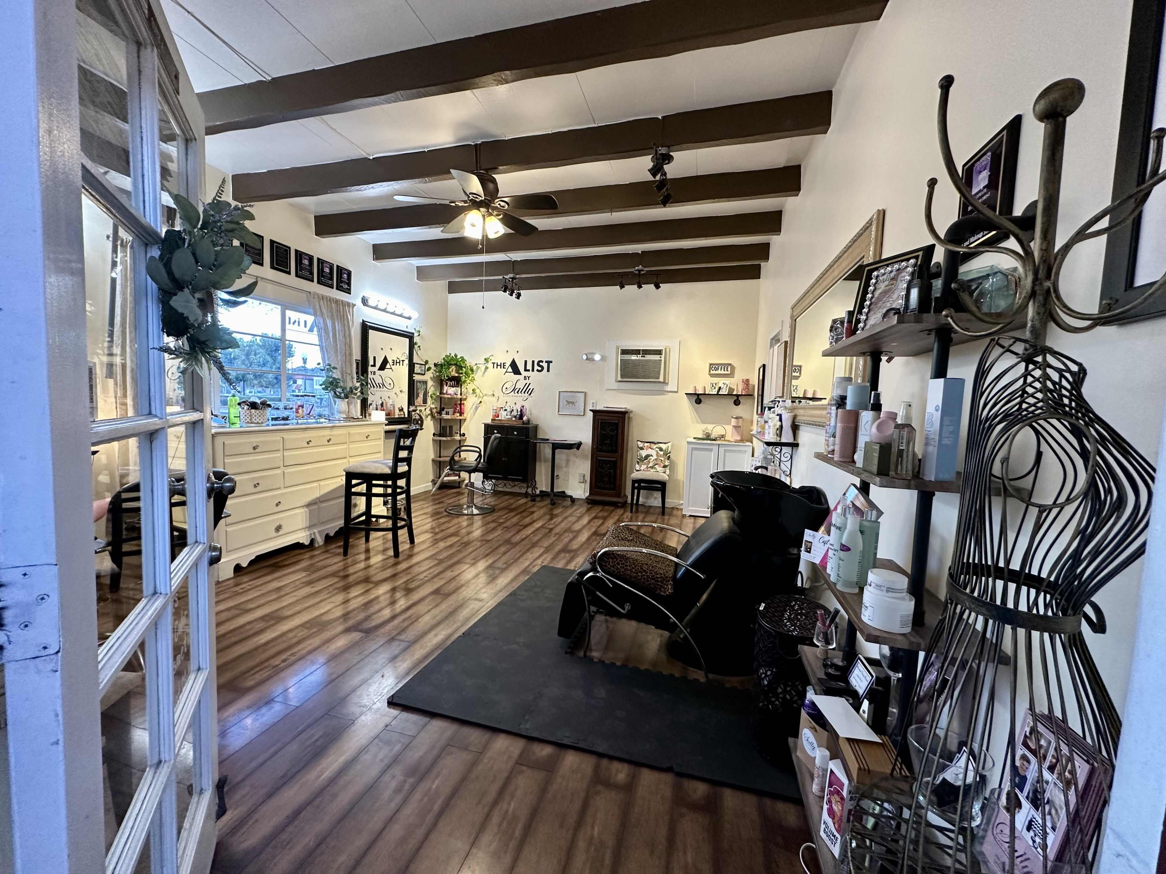 The image shows a hair salon with wooden beams on the ceiling, a waiting area with a black chair, and various styling stations arranged along the walls.