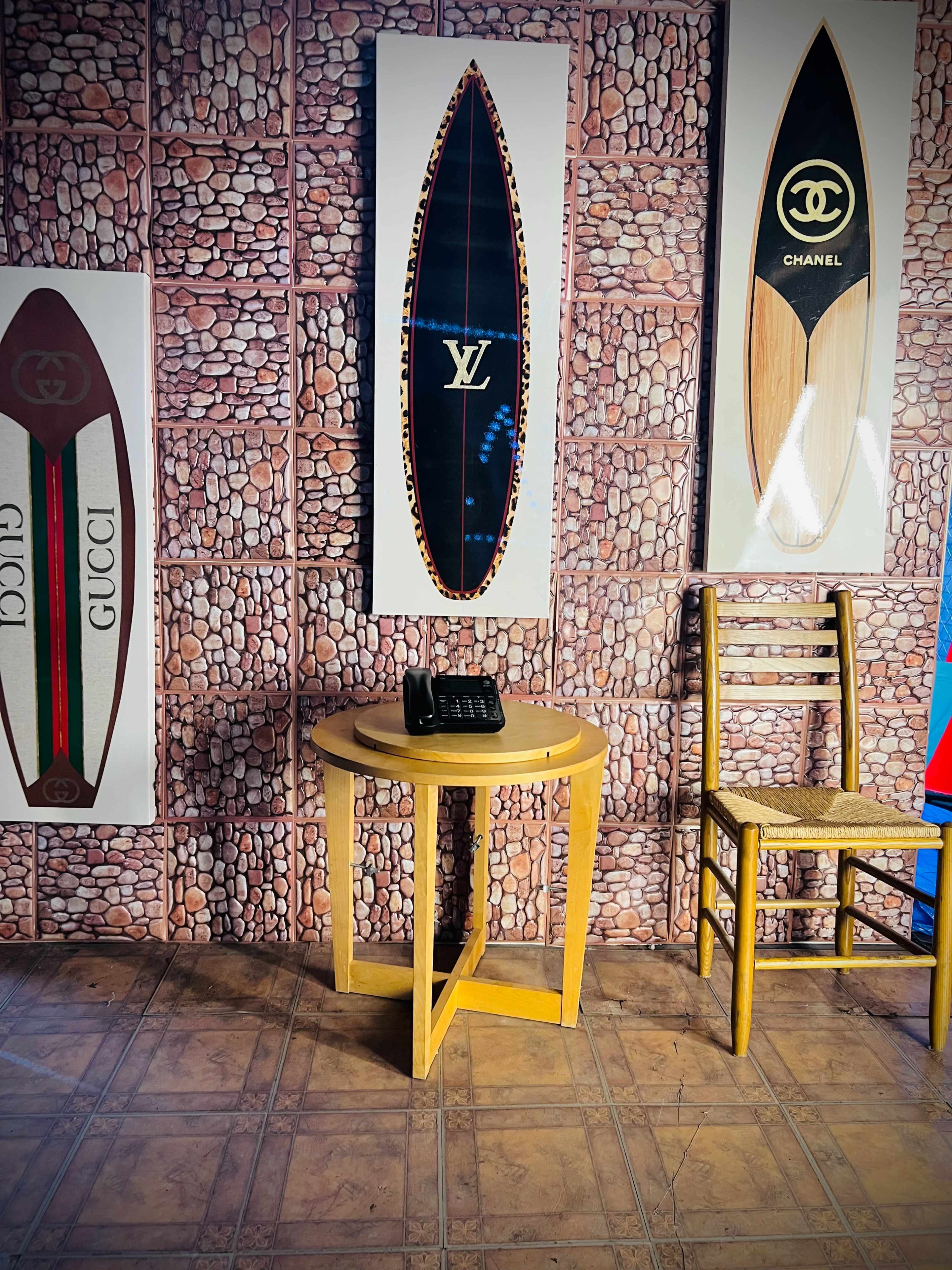The image shows a small wooden table with a phone on it beside a chair, against a wall decorated with surfboard art featuring luxury brand logos.