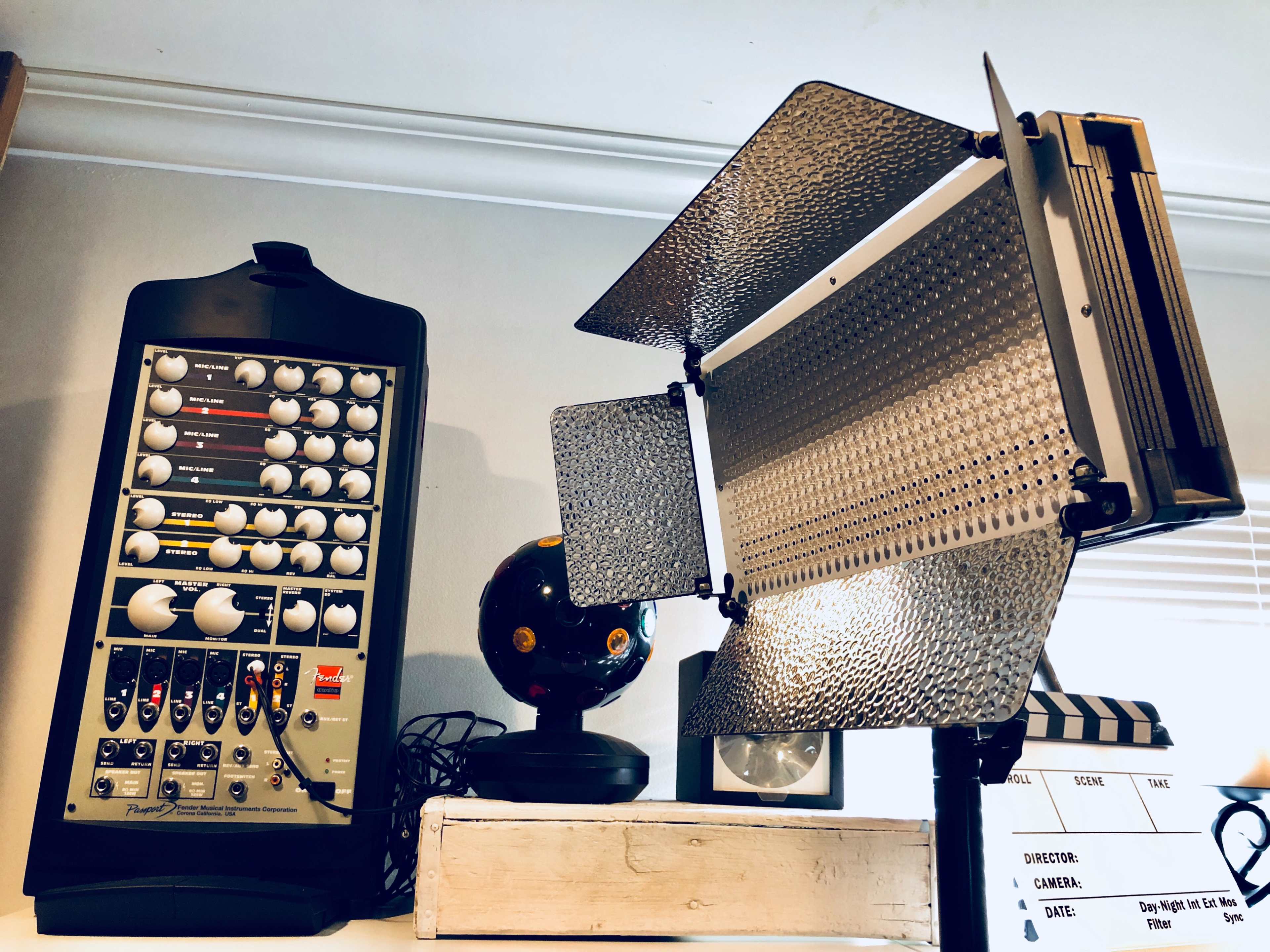 A vintage audio mixer next to a studio light with barn doors, placed on a shelf alongside a small globe and a movie clapperboard.