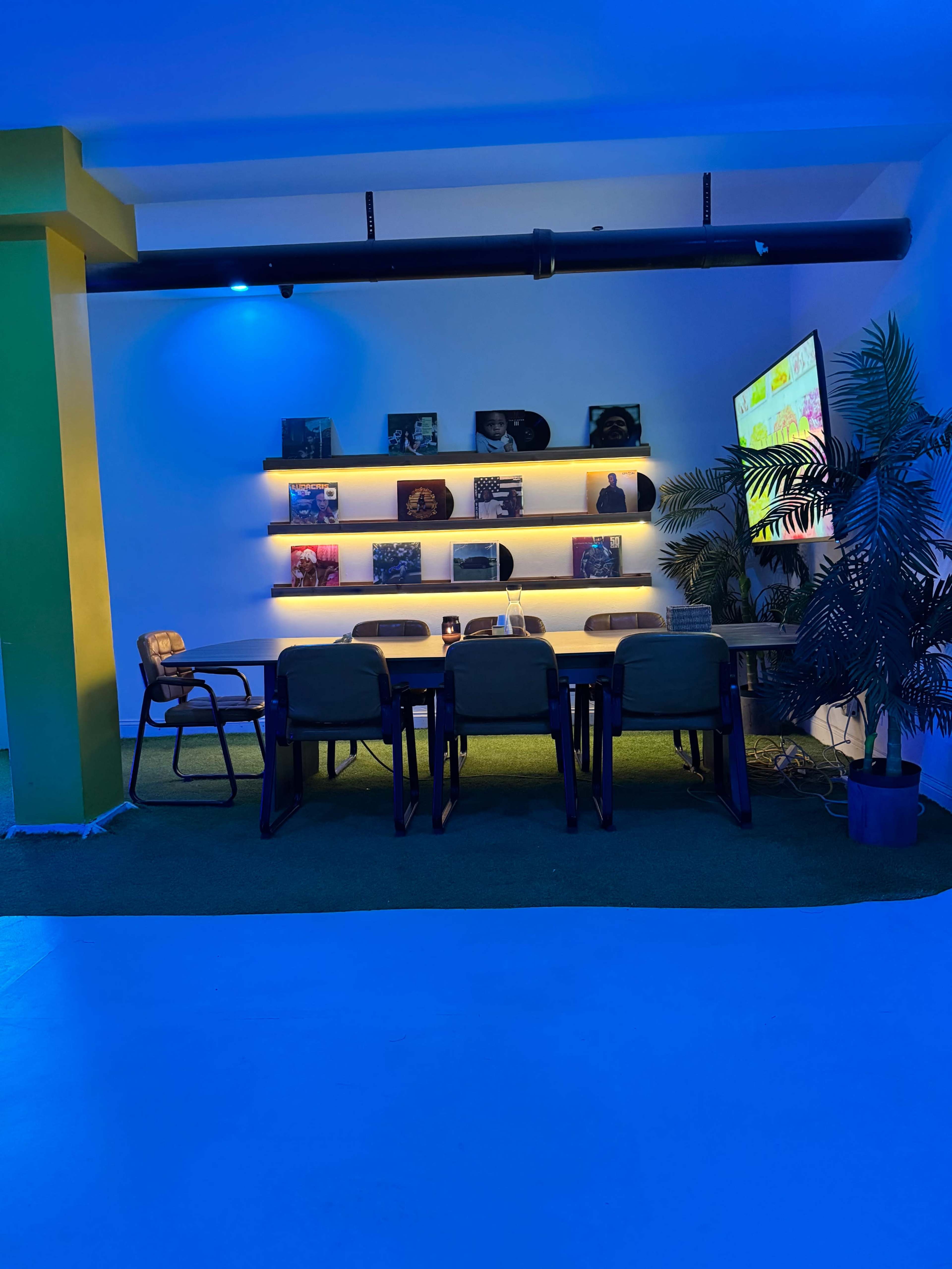 A brightly lit modern meeting room features a large table surrounded by chairs, with shelves displaying records and a television mounted on the wall.