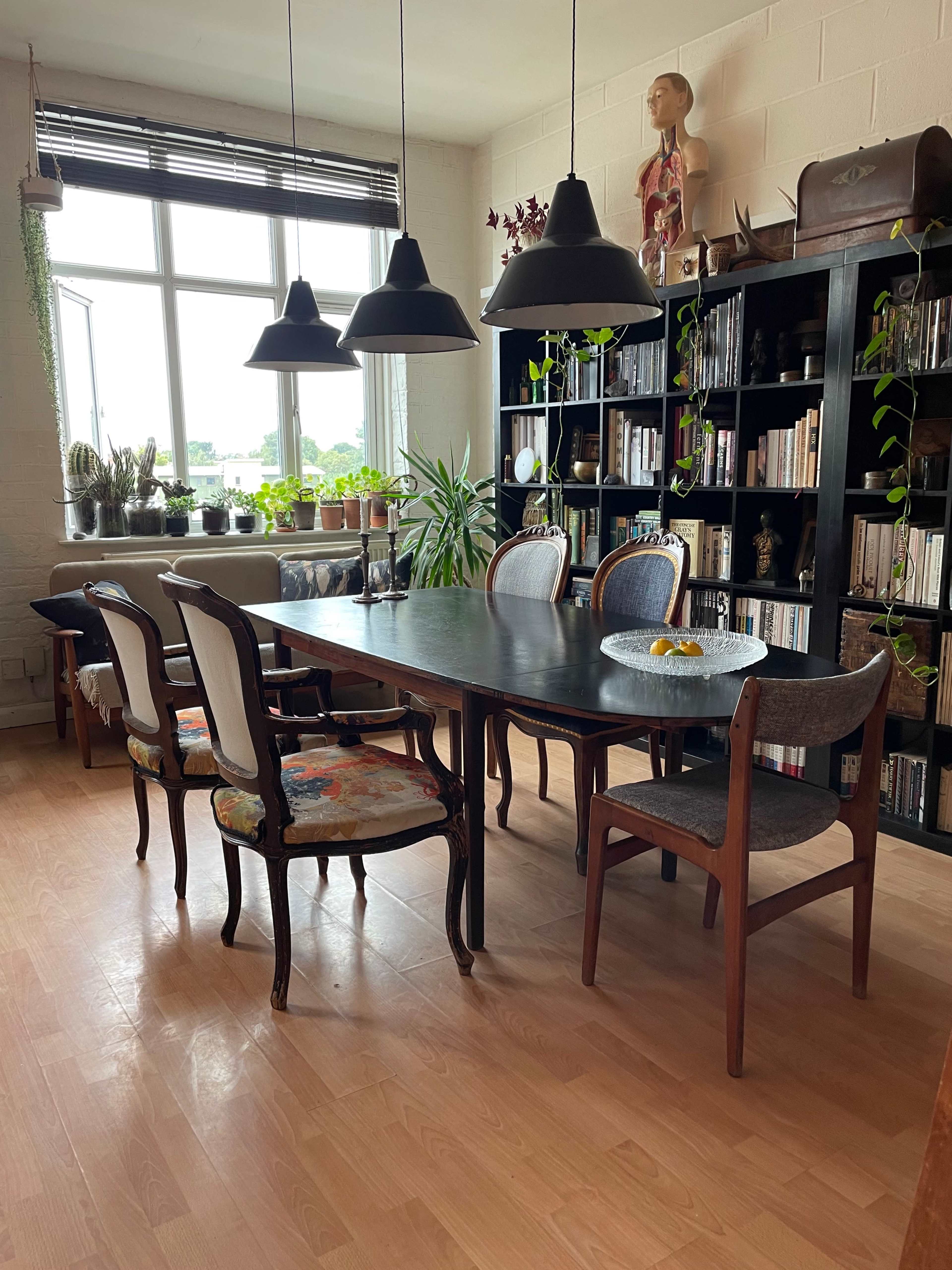 A dining table with six chairs is positioned in a well-lit room, accompanied by hanging pendant lights and a large bookshelf filled with books and plants.