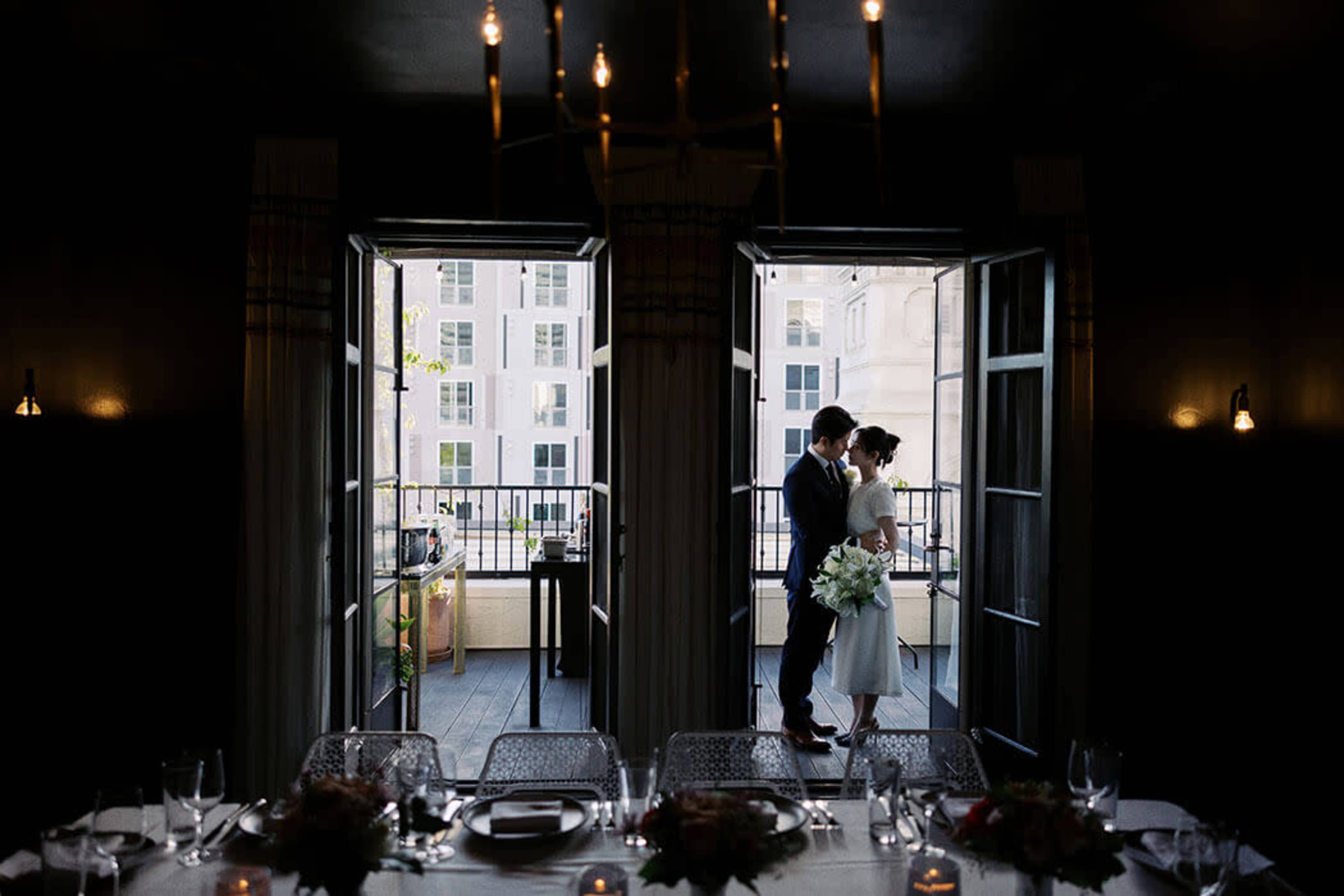 A couple stands together at an open balcony door, framed by large windows, while a dining table is set for a gathering in the foreground.