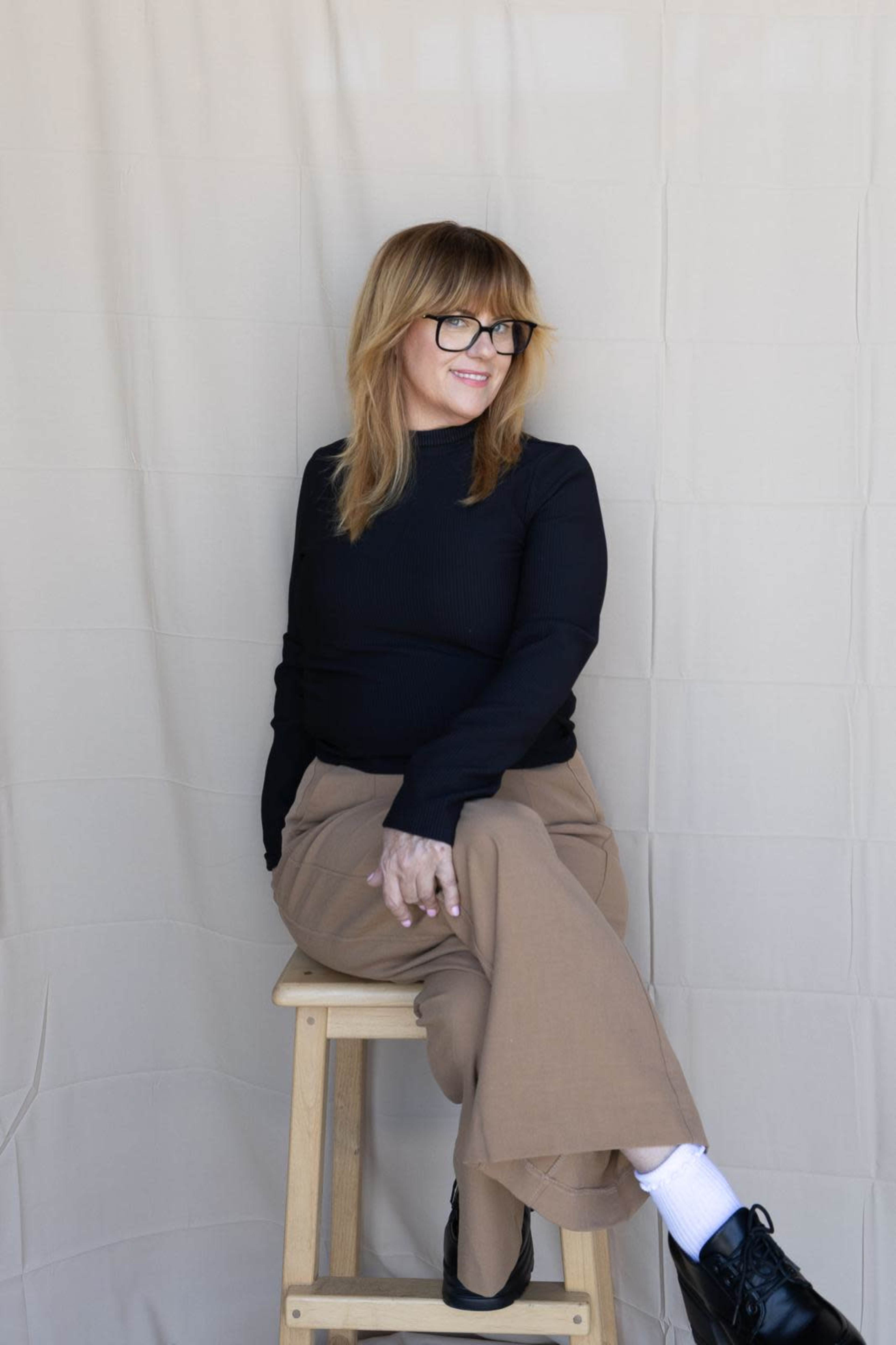 A person with glasses sits on a stool against a beige backdrop, wearing a black top and tan trousers.
