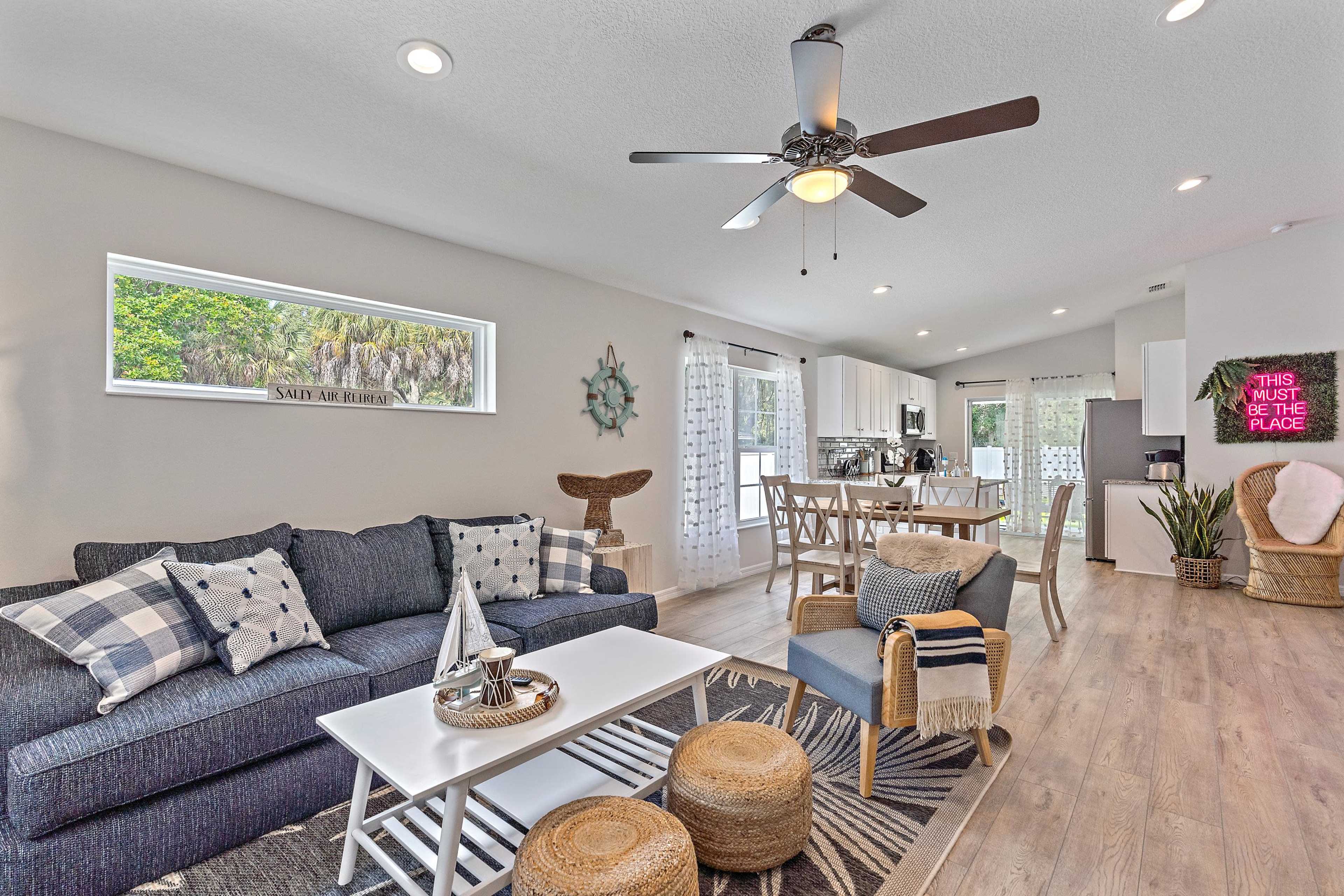 A modern living room features a dark blue sofa, a light coffee table, and natural fiber stools, with an open kitchen visible in the background.
