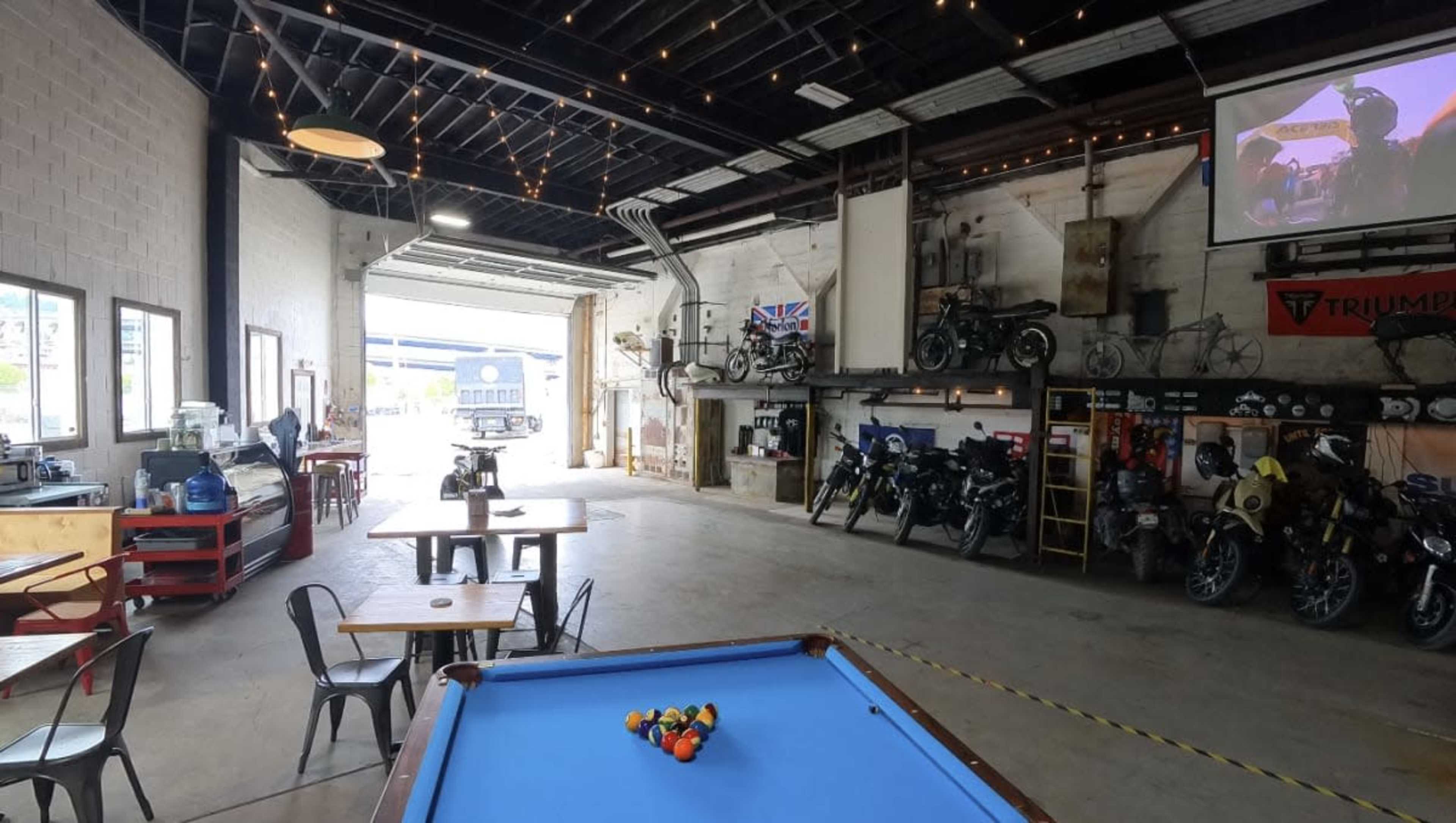 The image shows a spacious garage-like interior with a pool table in the foreground, motorcycle displays along the walls, and large windows opening to an outdoor area.