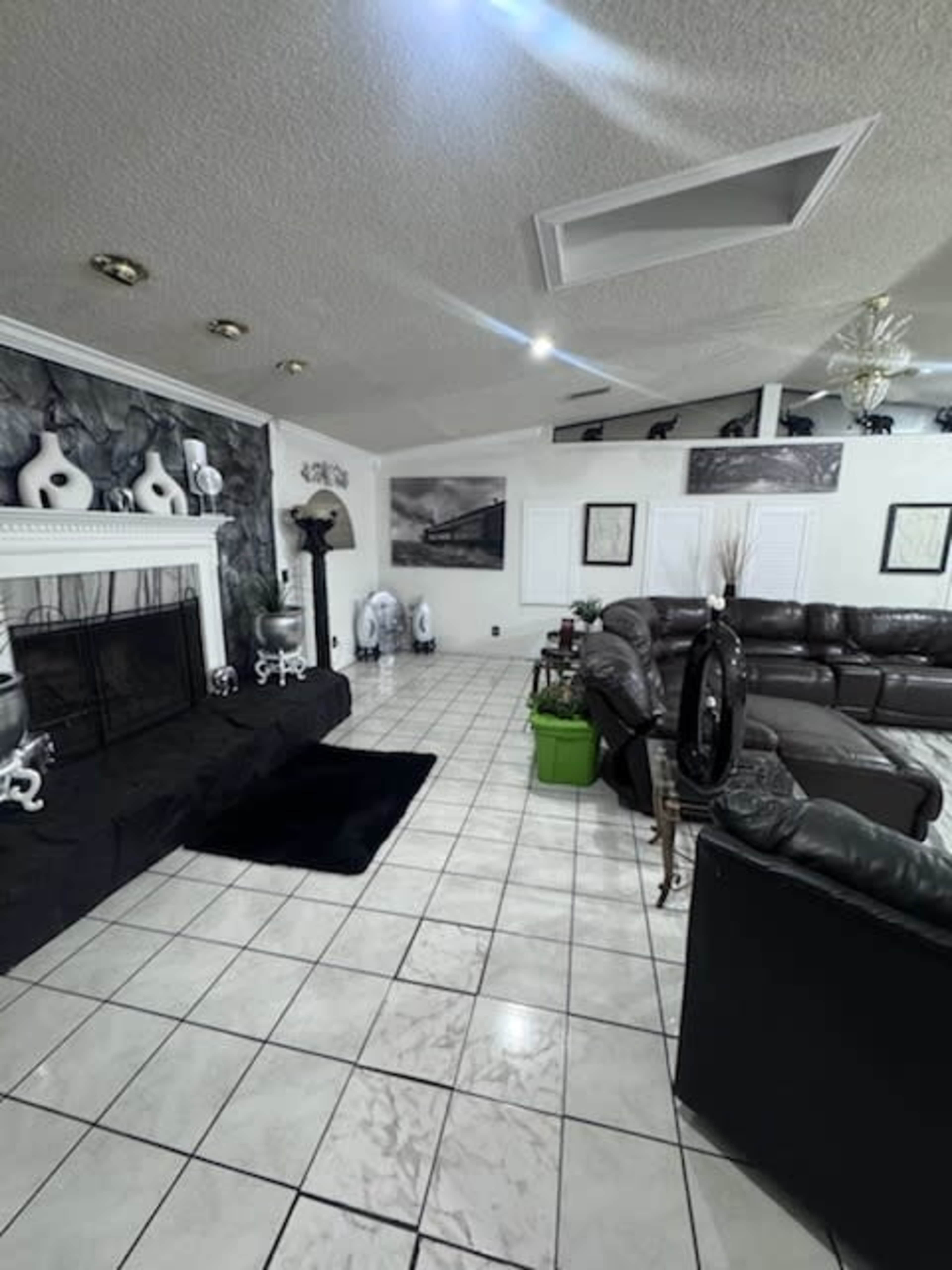 The image shows a spacious living room featuring a black sectional sofa, a decorative fireplace, and a tiled floor with various decorative elements and plants.