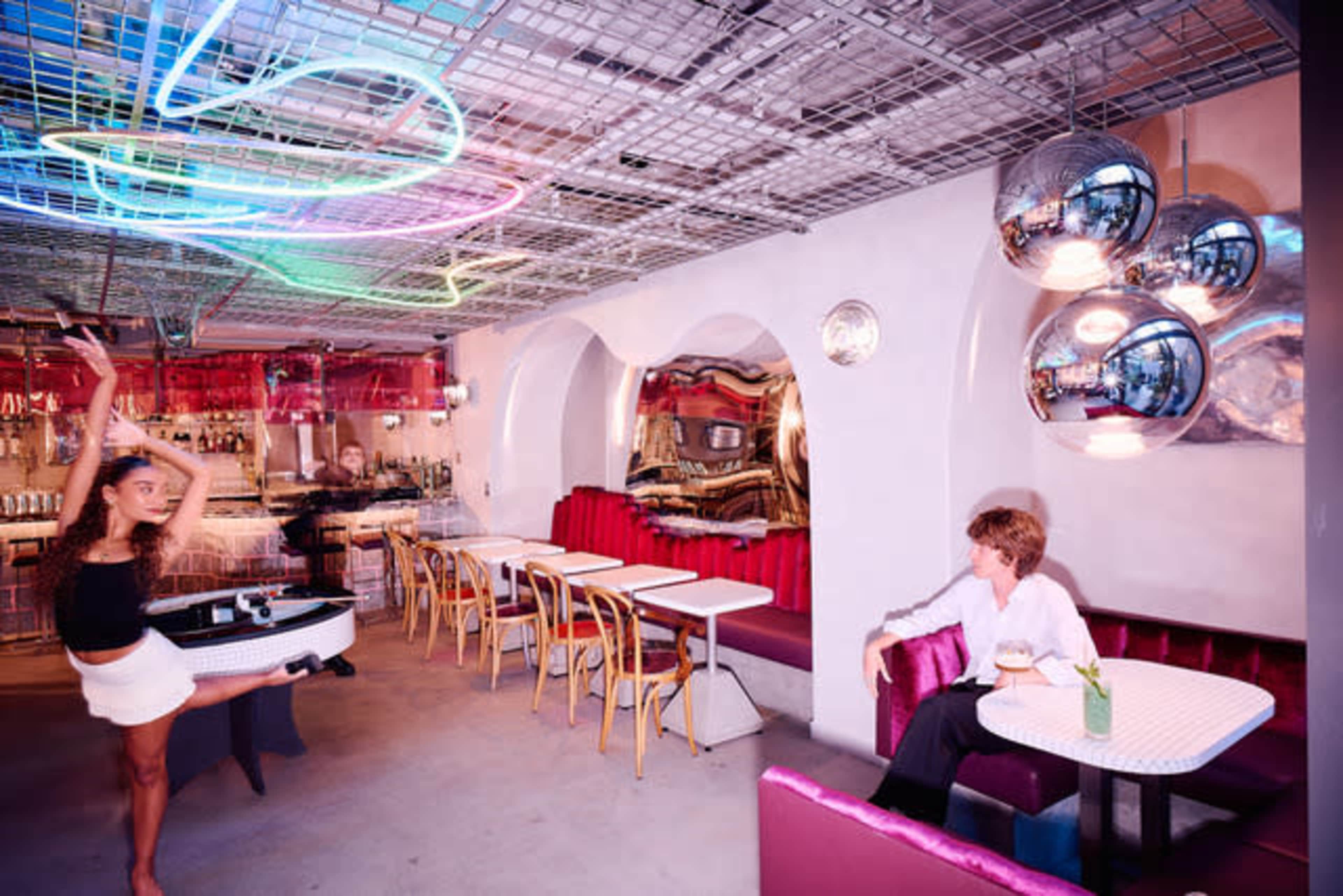 A dancer performs near a table in a modern restaurant featuring colorful neon lights and shiny decor.