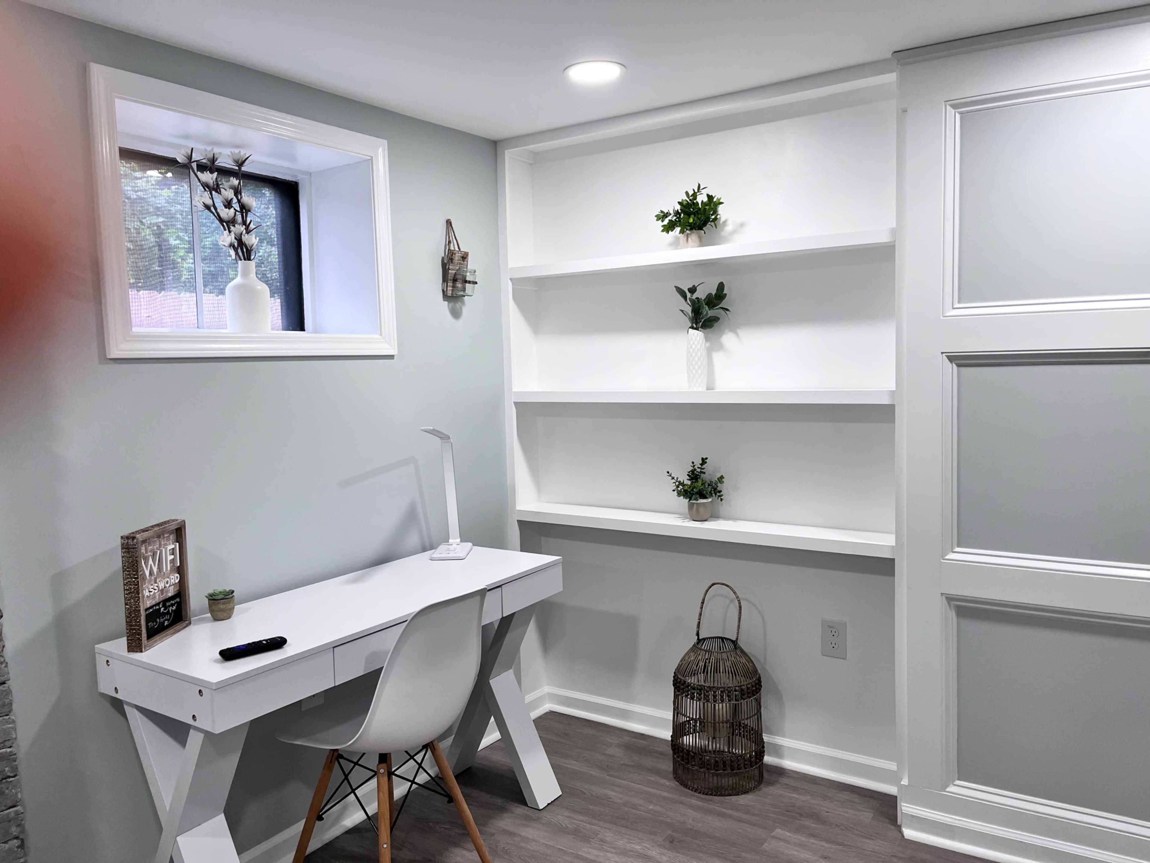 A small home office area featuring a white desk, shelves with decorative plants, and a window.
