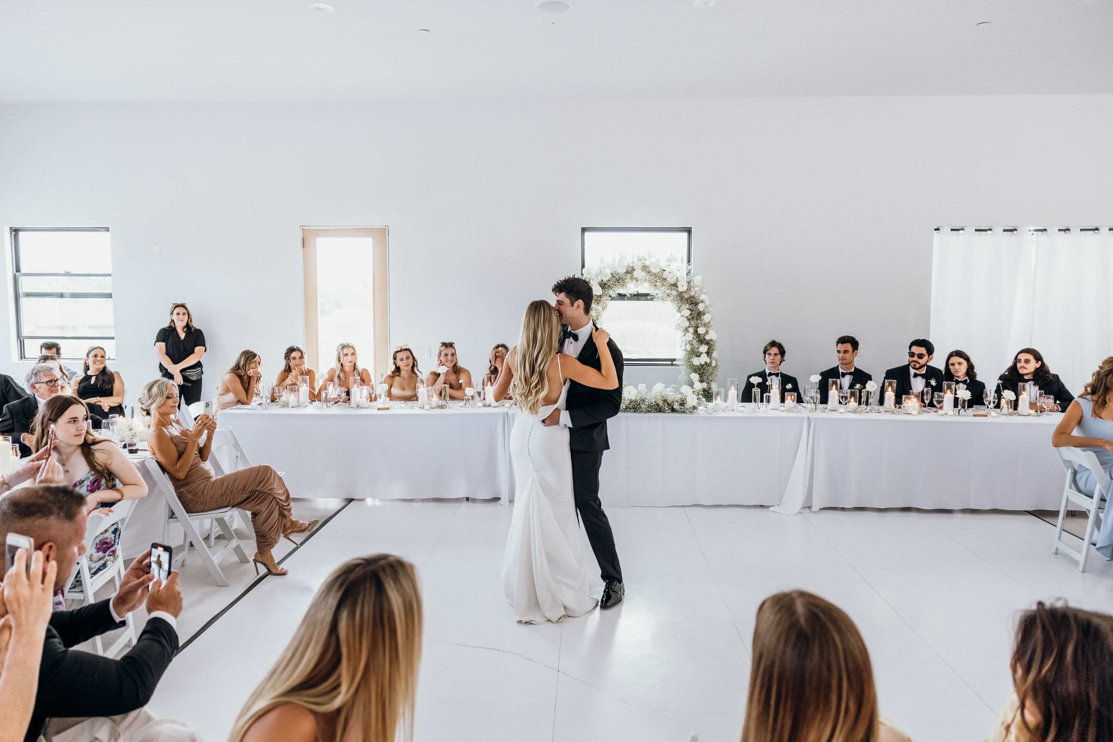 A couple dances together at their wedding reception while guests watch and sit at a long table adorned with candles and decorations.