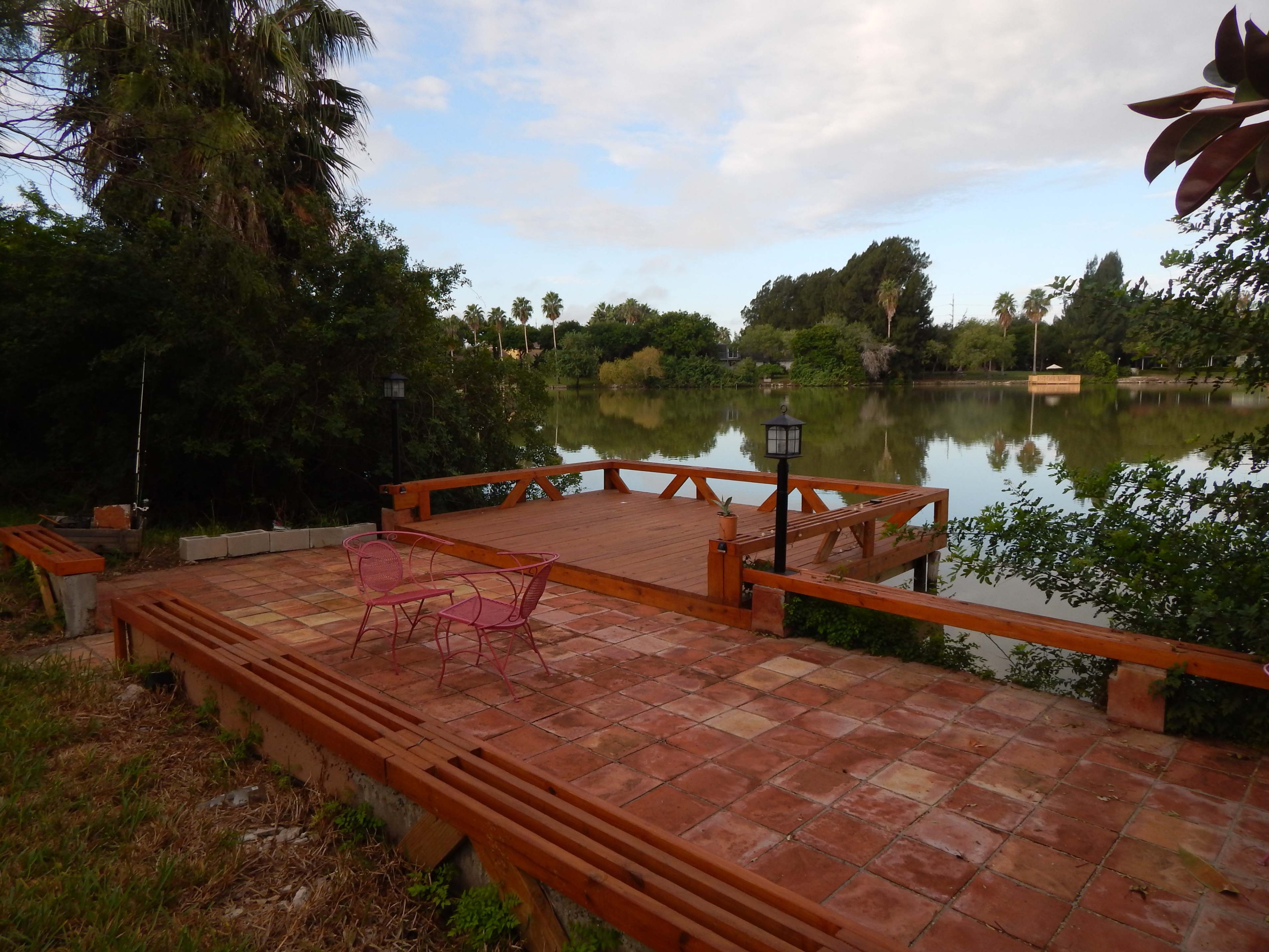 A wooden deck with pink chairs overlooks a calm lake surrounded by greenery and palm trees.