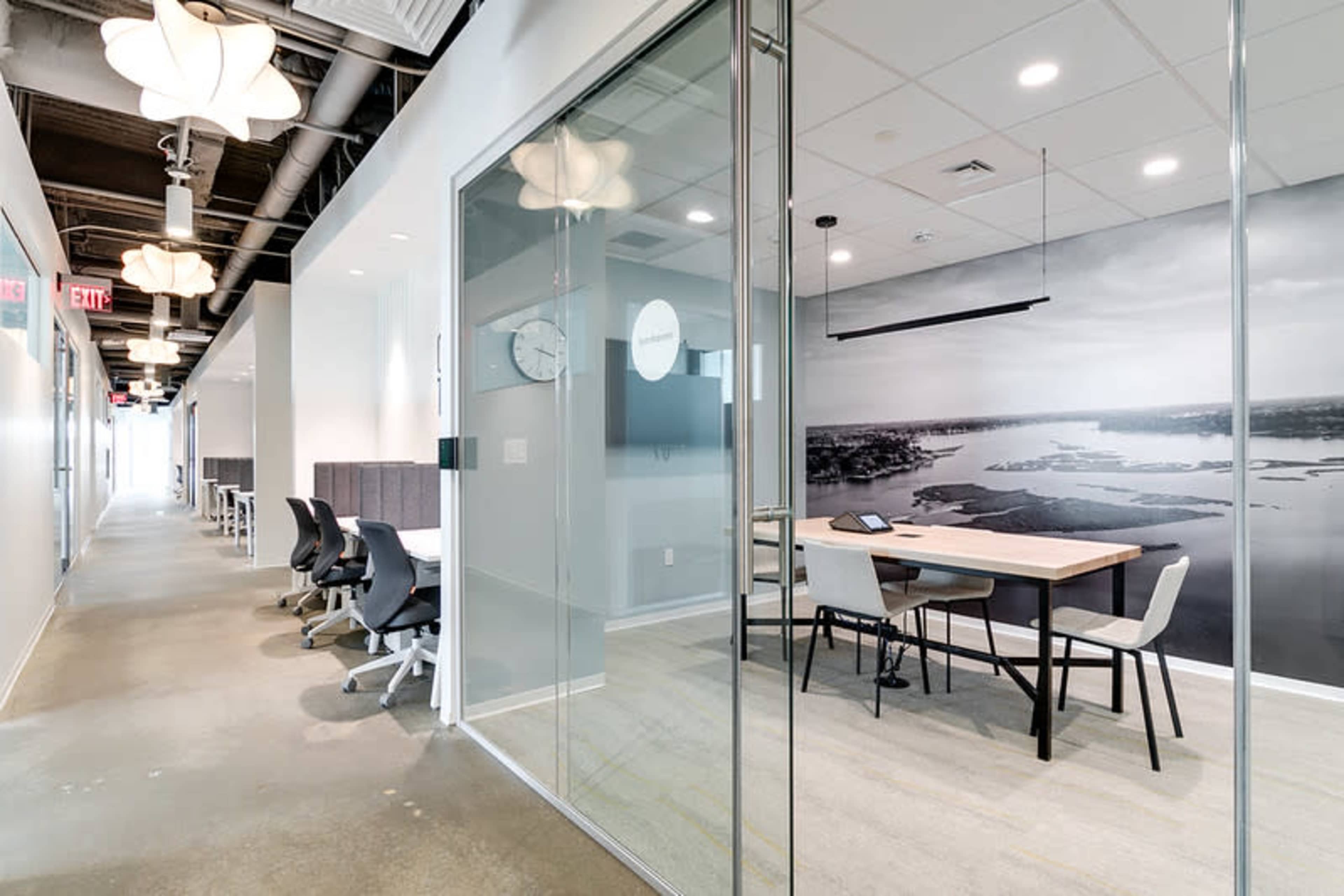 The image shows a modern office space featuring a glass-walled conference room with a wooden table and chairs, adjacent to a hallway lined with workstation cubicles.