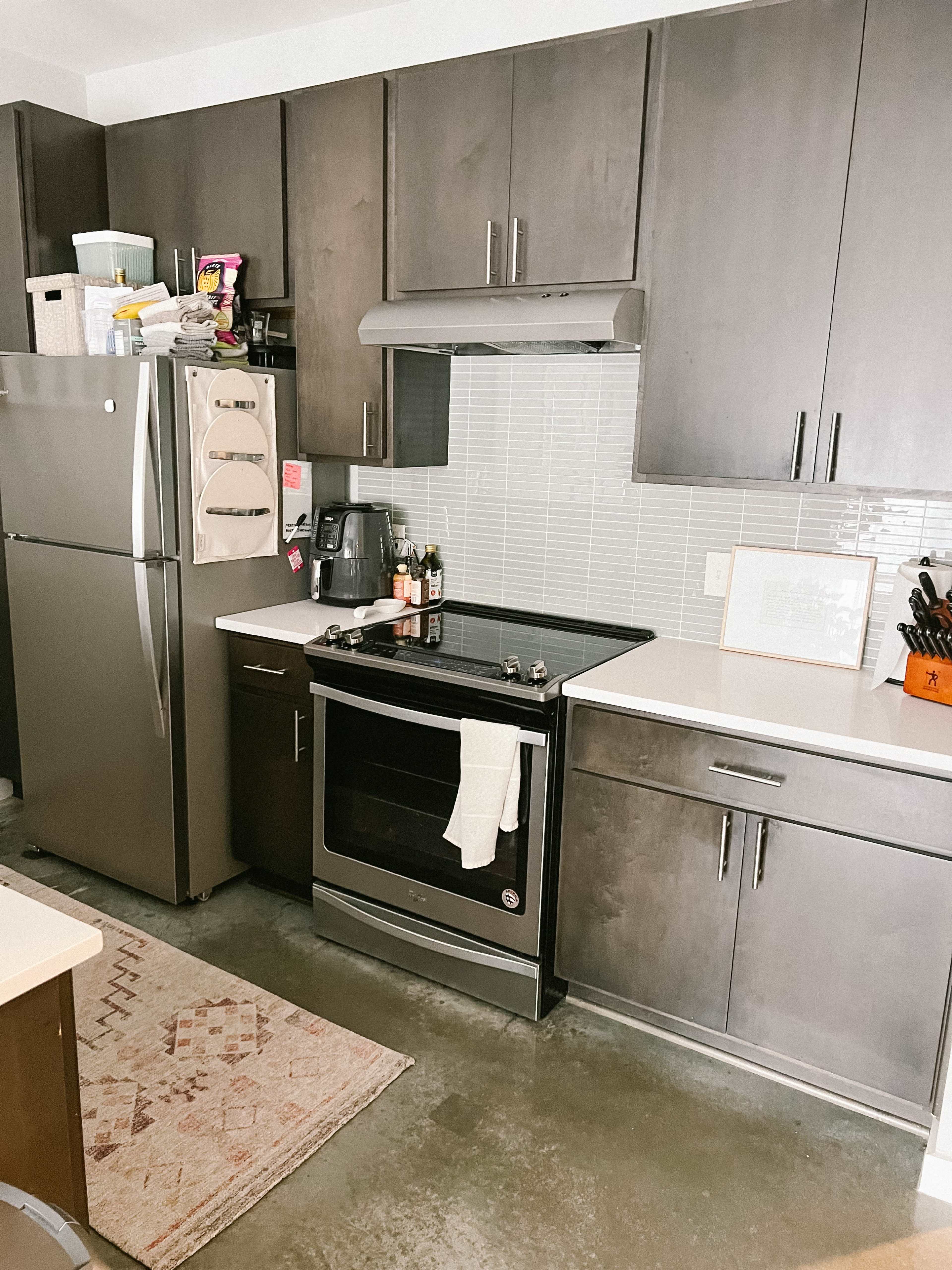 A compact kitchen features stainless steel appliances, dark cabinets, and a white countertop with a rug on the floor.