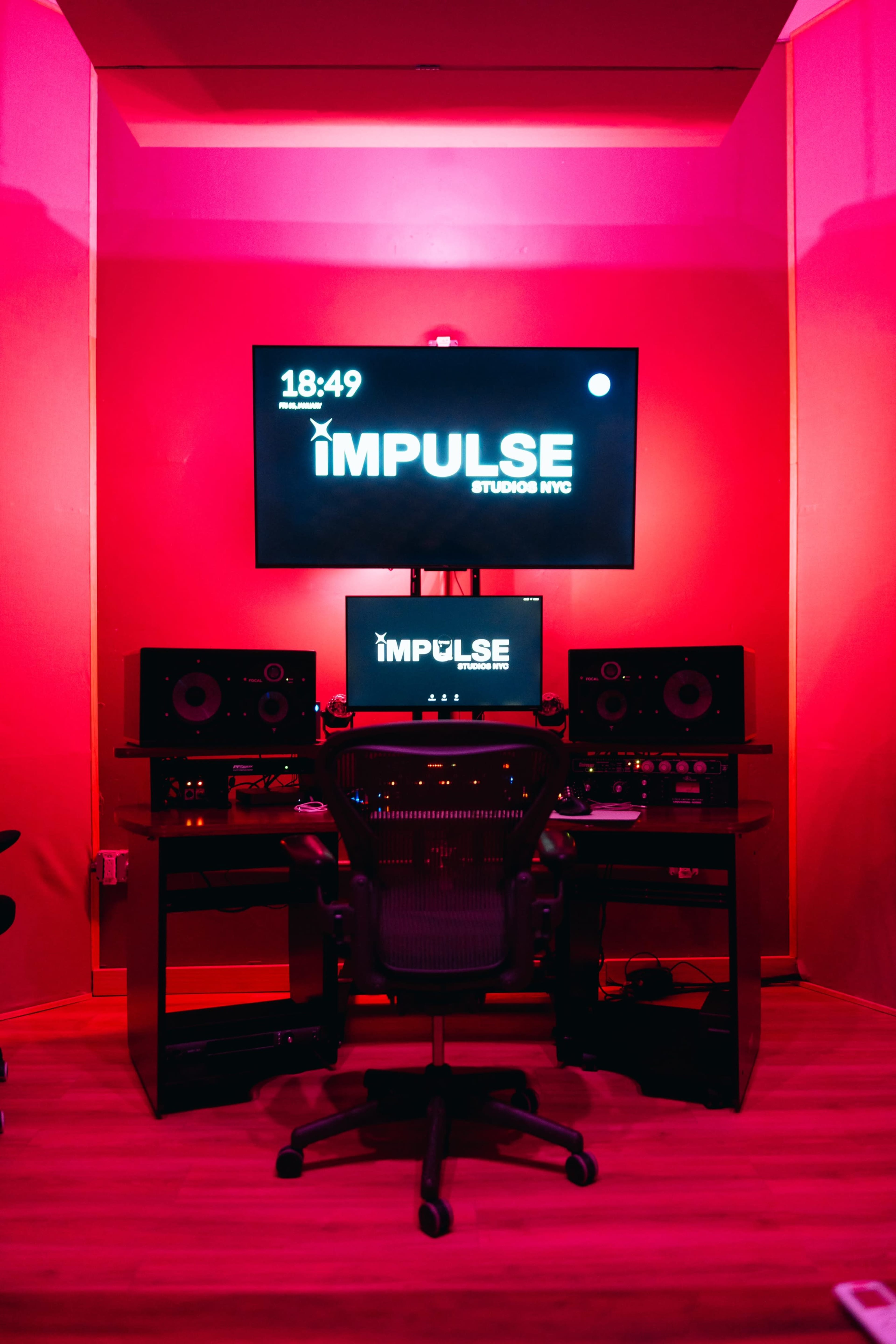 A music production studio is illuminated with pink lights, featuring a desk with two monitors, speakers, and a black ergonomic chair.