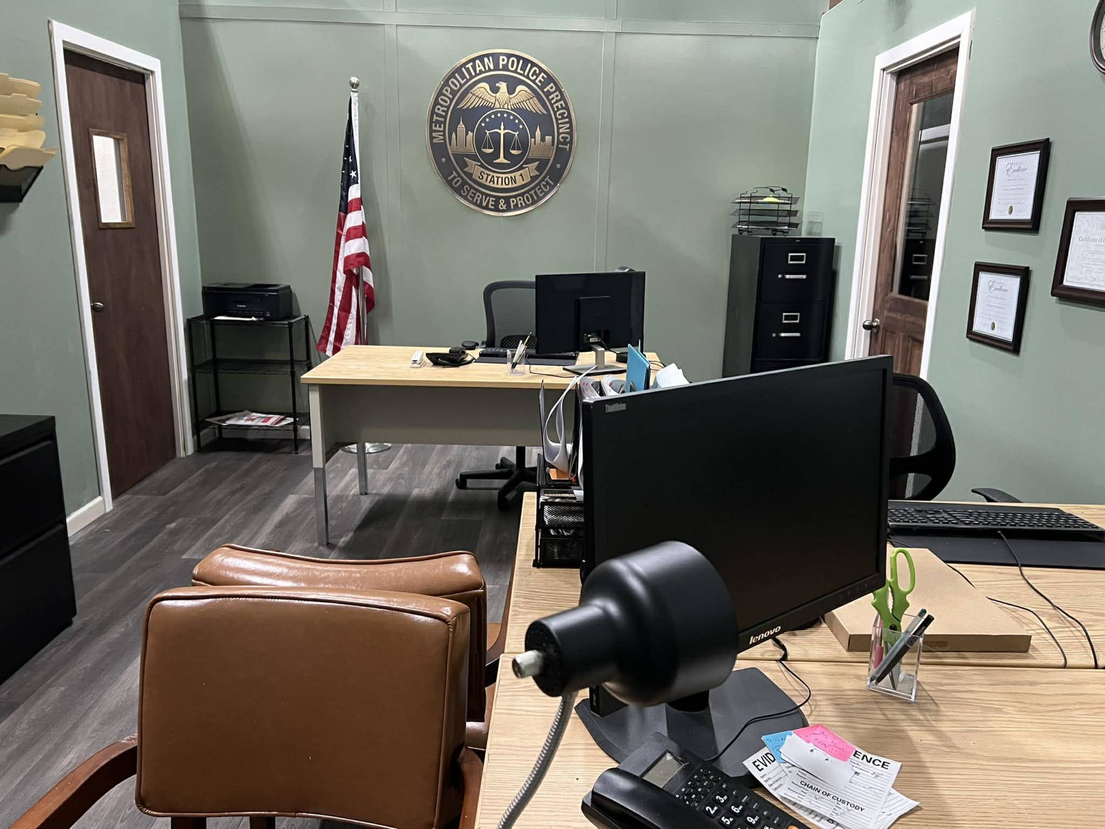 The image shows a small office space featuring two desks, a computer setup, an American flag, and a wall-mounted emblem of a police department.
